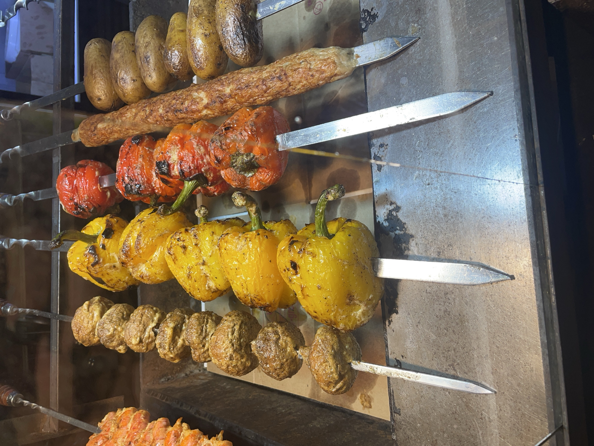 The image shows skewers of food being cooked in a restaurant kitchen. On the left, there are skewers of what appear to be meatballs or falafel, alternating with yellow bell peppers. Next to them are skewers of red bell peppers and a long, cylindrical skewer of ground meat, possibly kebab. To the far right are skewers of small, whole potatoes. The food is grilled and has visible char marks. The skewers are resting on a metal surface, with a checkered paper liner underneath. The background shows a stainless steel shelving unit and a hint of boxes. The lighting suggests overhead cooking lights.