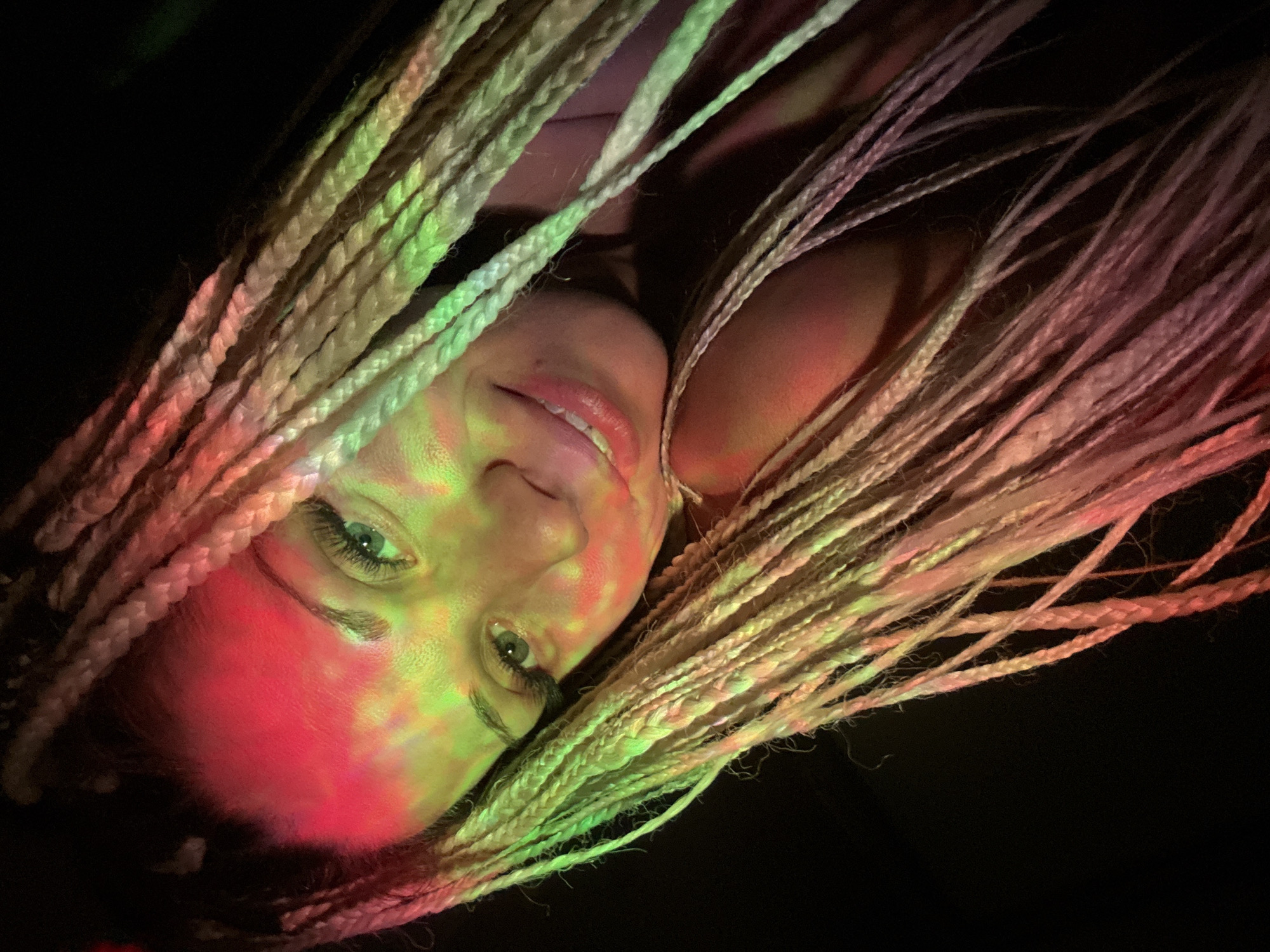The image is a close-up, low-angle shot of a young adult female with blonde braided hair. She is lit by colorful lights, with red and green hues prominently displayed on her forehead and face. Her eyes are green and appear to be looking towards the camera with a slight smile. Her hair is long and styled in numerous braids that fall around her face and shoulders. The background is dark, suggesting an indoor setting with artificial lighting. The photograph does not contain any recognizable landmarks or environmental clues that would indicate a specific location within the city of Rusanovo, Russia.