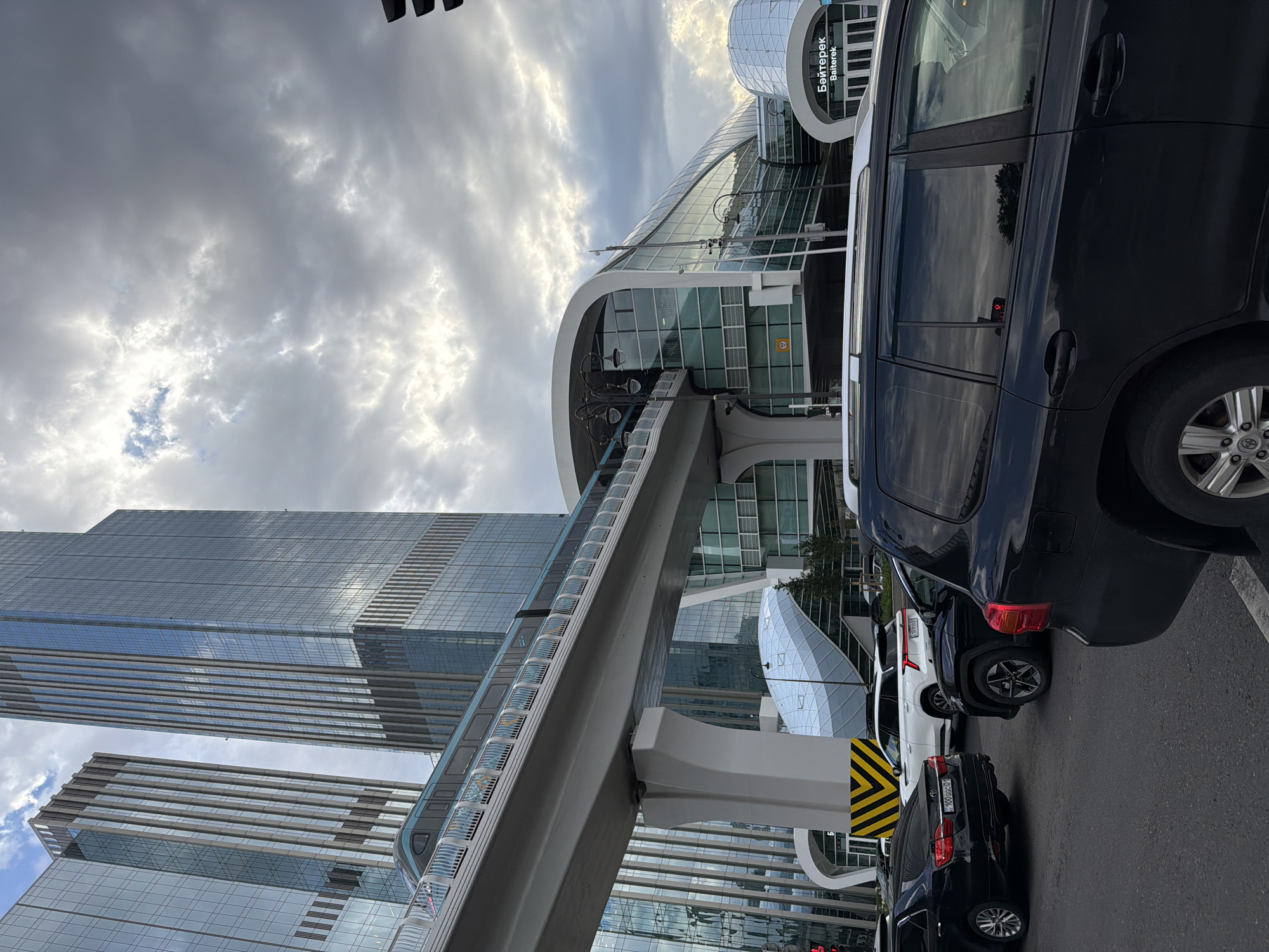 An exterior, eye-level, medium shot captures a contemporary urban scene in Astana, Kazakhstan, under a cloudy sky. In the foreground, several dark-colored vehicles are stopped at an intersection, suggesting traffic. Behind them, a modern elevated monorail train glides along a sleek, white track that passes in front of a striking building with a curved, glass facade. The building is part of a larger complex, with other contemporary architectural structures, including one with a domed roof. Text in Cyrillic and Latin script, reading "Байтерек Baiterek," is visible on a sign within the complex, indicating this is the Baiterek area of Astana. Two tall, modern glass skyscrapers dominate the background, reflecting the cloudy sky.