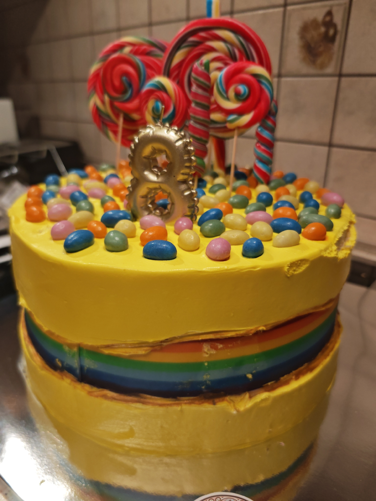 A yellow frosted cake with a rainbow-colored layer is decorated with colorful candies and lollipops. A golden numeral "8" candle sits on top of the cake, and three spiral lollipops are placed behind it. The cake is presented on a silver cake board. The background consists of a tiled wall. The image was taken indoors.