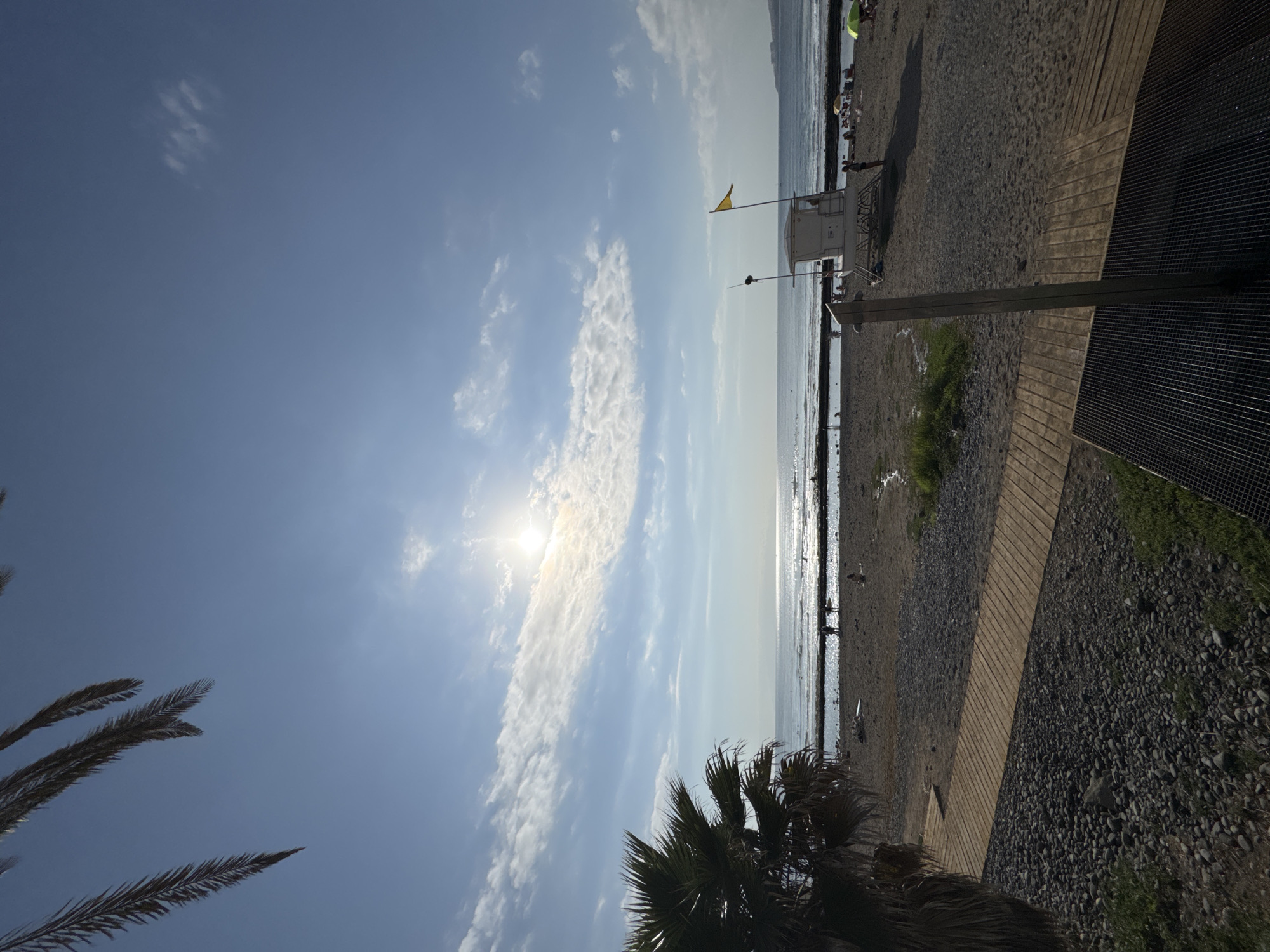 The image depicts a sunny day at a beach in Arona, Spain. In the foreground, a wooden walkway leads towards the sandy beach, with a metal grate structure, likely an outdoor shower, visible on the right. To the left, a palm tree's fronds hang over the scene, with pebbles and sparse green vegetation occupying the immediate foreground. The beach itself is populated with a few individuals, some walking near the water's edge and others relaxing on the sand. A lifeguard station, complete with a yellow flag, stands prominently on the beach. The ocean stretches out to the horizon, reflecting the bright sun, with a faint line of clouds across the clear blue sky. The overall atmosphere is one of a relaxed, seaside environment.