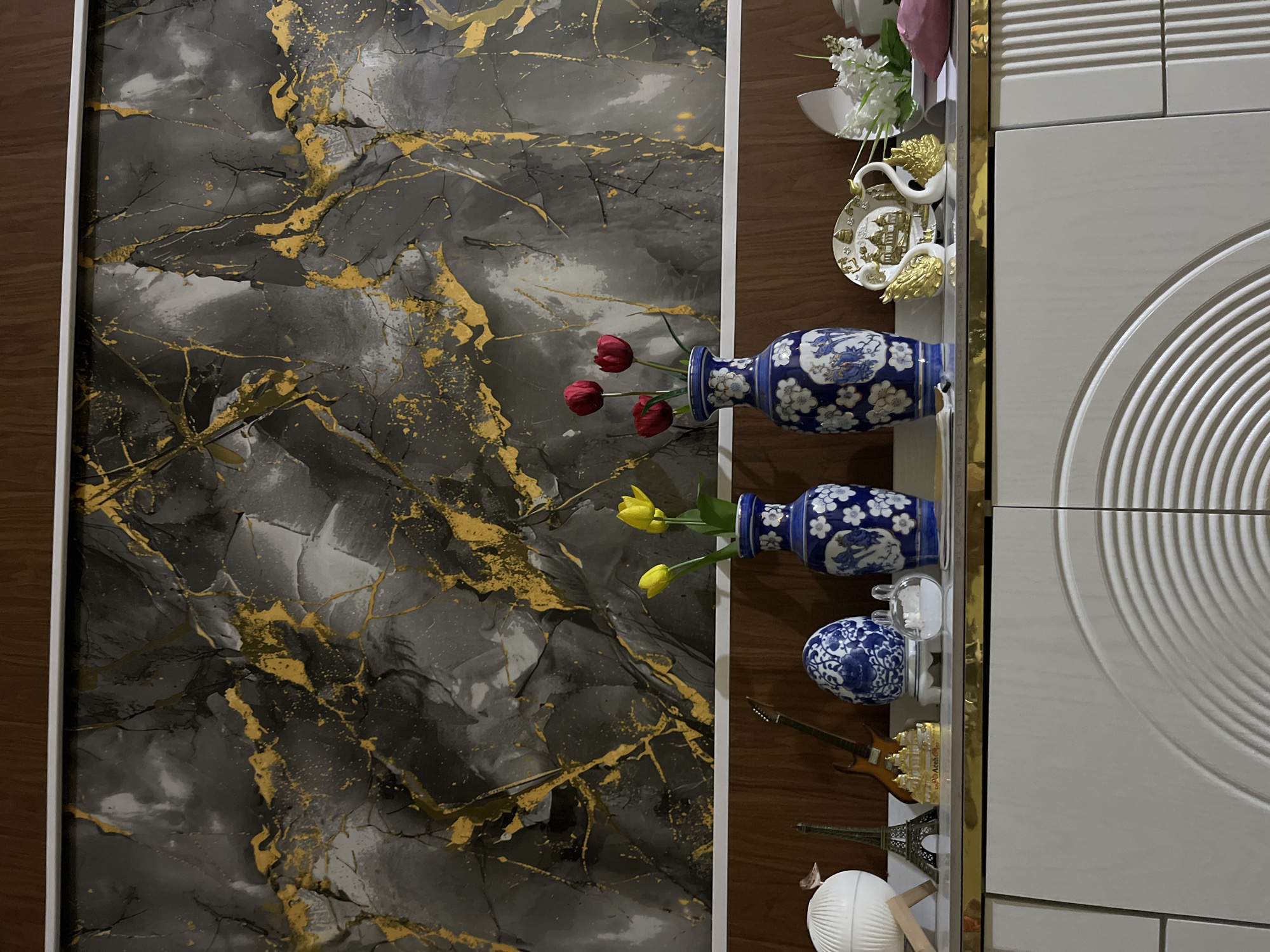 The image showcases an interior decorative display, likely a shelf or console positioned against a prominent feature wall. The main backdrop on the left is a large panel adorned with an abstract, marbled design, featuring dark gray and black tones interlaced with striking golden streaks and splatters, suggesting a luxurious stone or artistic finish. This panel is bordered by a thin white frame and a thicker dark wooden frame.
To the right, a wooden shelf or console runs vertically, holding an assortment of decorative objects. Along its right edge, the shelf is highlighted by a narrow, shiny gold trim. From top to bottom, the items include a small white decorative plate with gold, possibly winged or angelic, embellishments. Below it, a blue and white patterned ceramic vase with floral and bird motifs contains three artificial red tulip-like flowers. Further down, another similar blue and white ceramic vase holds two artificial yellow tulip-like flowers. A smaller, rounder blue and white ceramic decorative object is placed nearby.
Towards the lower section of the shelf, miniature figurines are visible: a brown electric guitar and a golden Eiffel Tower replica. At the bottom, a white spherical object, resembling a paper lantern or decorative orb, is present. Artificial white flowers and a touch of pink fabric are also visible among the upper decorations.
To the far right, a light-colored wall or cabinet features architectural detailing, including horizontal ridged lines at the top and a distinctive large circular pattern composed of concentric rings on its lower part. A vertical gold trim separates this section from the wooden shelf area. The setting appears to be indoors, under soft, even lighting. There are no people or activities depicted, and no visible text.