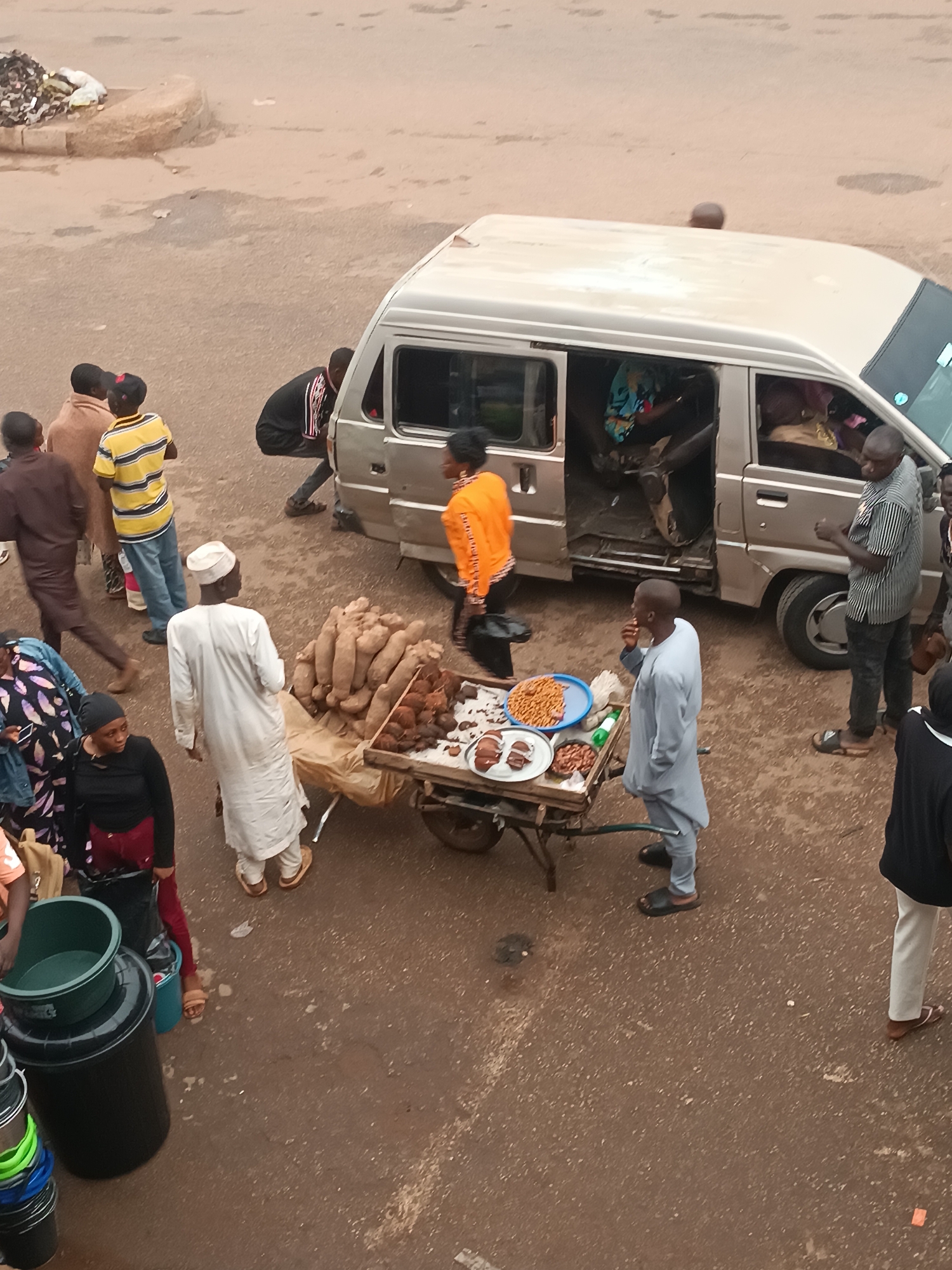 The image depicts a street scene in an urban environment. A silver minivan is parked on the right side of the frame, its side door open, with several people interacting with it. A person in an orange shirt is exiting the vehicle. Several individuals are standing near the minivan, including one in a black and white striped shirt.
In the foreground, a street vendor is positioned with a pushcart laden with goods. The cart displays items such as yam tubers and various processed foods. A person dressed in white is examining the items on the cart. Another person, dressed in light blue, is standing near the cart and seems to be eating.
Several other individuals are milling around the area, some walking and others standing. Various containers, including buckets, are clustered on the lower-left side of the frame. The overall setting appears to be a busy street market area.