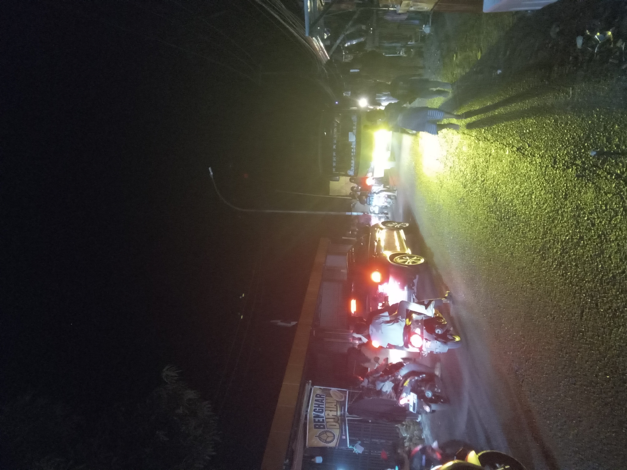 This is a night-time, low-light, street-level, vertical shot of a busy road in an urban environment. The road is paved and appears wet, reflecting the bright yellow headlights of a vehicle and the red taillights of other vehicles. Several people are on motorcycles, some of whom appear to be riding in traffic, while others are stationary. There are buildings along the side of the road, including one with a banner that reads "BENGHAR DURIAN". A street light is visible in the distance, illuminating a portion of the sky. There are also overhead wires and poles, indicating the presence of infrastructure. The overall atmosphere is one of a lively, active street scene at night.