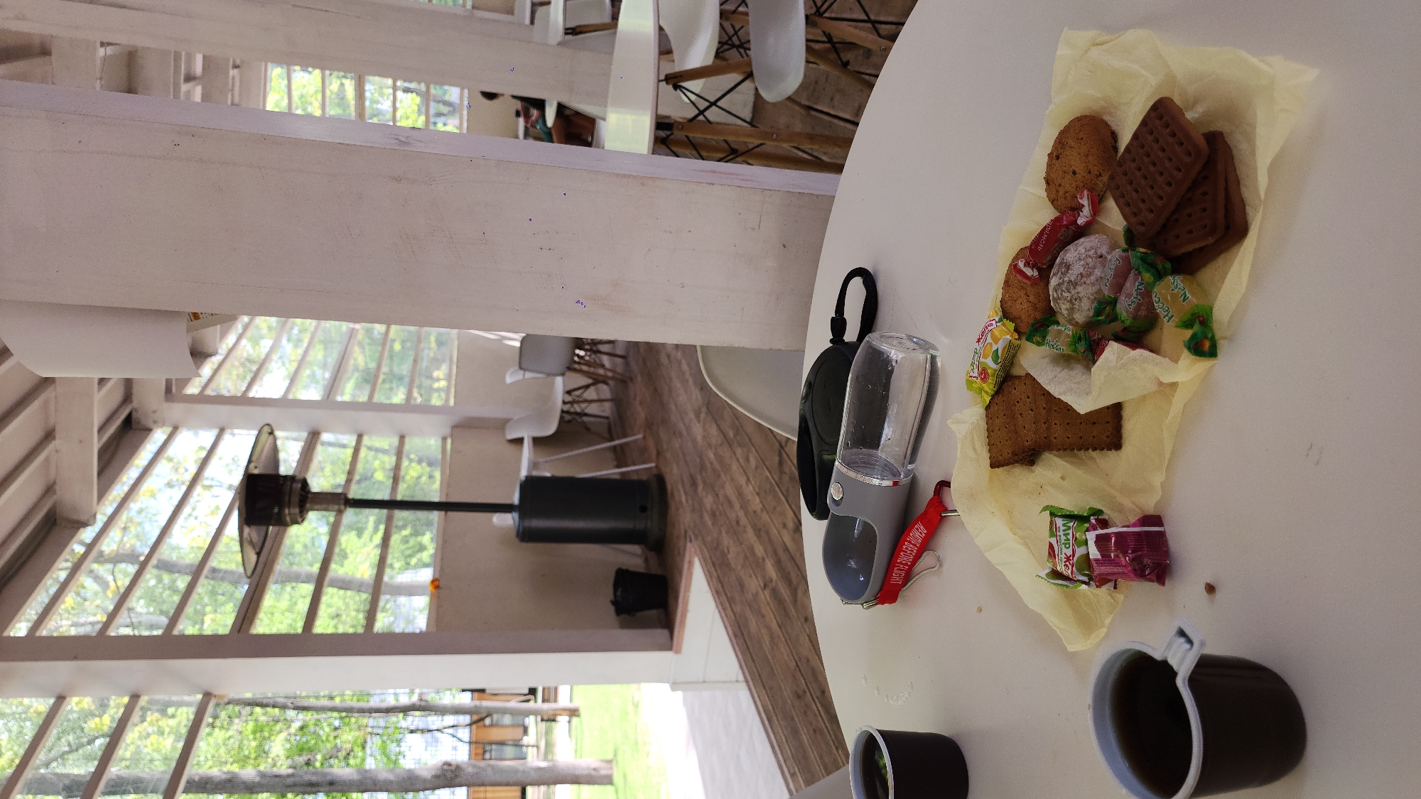 On a white table, likely outdoors given the visible building structure and greenery, is a collection of items: a small portable dog water bottle with a black leash attachment, a scattering of cookies and candies on yellow paper, and two dark-colored mugs. The environment appears relaxed, possibly a patio or terrace. The presence of a patio heater suggests the location is intended for use even in cooler weather. This image does not show sufficient detail to confirm the location in the city of Khimki, Russia. There are no visible people.