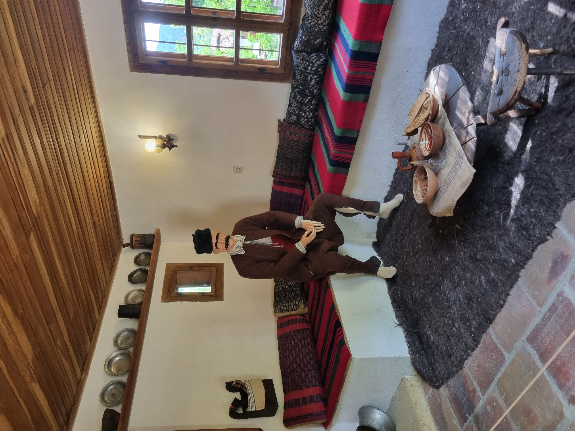 The image depicts an interior scene, possibly a museum exhibit, set in Elena, Bulgaria. A mannequin dressed in traditional Bulgarian attire, including a brown jacket, pants, and a black hat, sits on a built-in banquette covered with woven textiles in various patterns and colors. To the mannequin's right is a low wooden table with pottery and other objects arranged on top. A dark, roughly-woven rug covers the floor near the table.
In the background, a window with a wooden frame reveals an outdoor view of greenery. The walls are white, and a wooden ceiling is visible. On one wall, a collection of metal plates and other decorative items is displayed. A smaller, framed window is also present. Overall, the scene seems to represent a traditional Bulgarian home or setting, likely within the historic city of Elena.