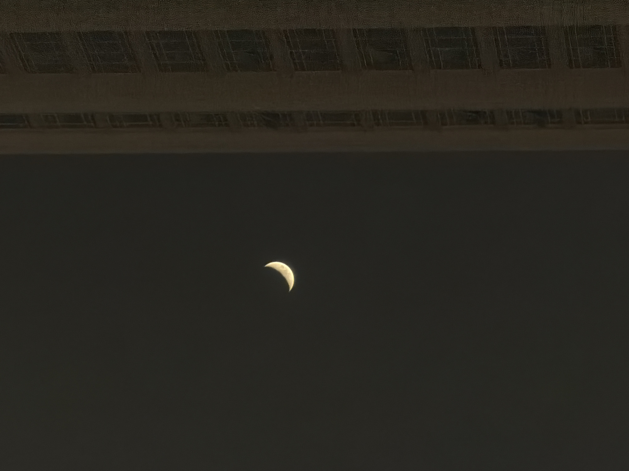 A crescent moon is visible in a dark sky to the left of a tall, light-colored building. The building has many windows, arranged in vertical rows. The scene appears to be in a city, though the specific location is unknown. The lighting suggests it is nighttime or dusk.