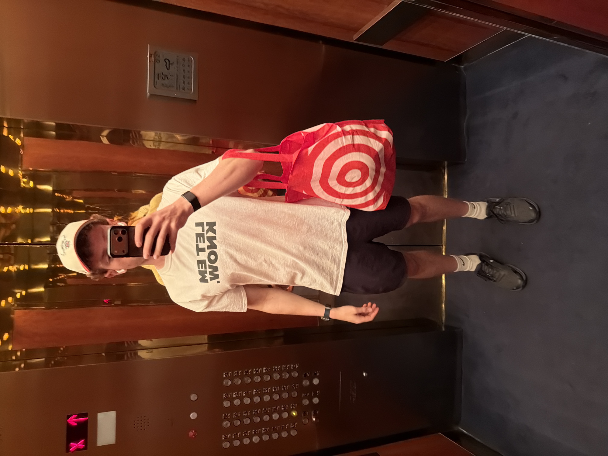 A person wearing a white T-shirt, shorts, and a baseball cap takes a selfie in an elevator. The person is also carrying a red Target shopping bag. The elevator has a panel with multiple buttons and a directory. The elevator appears to have wood paneling and is located in the United States of America. No further specifics of this location can be determined.