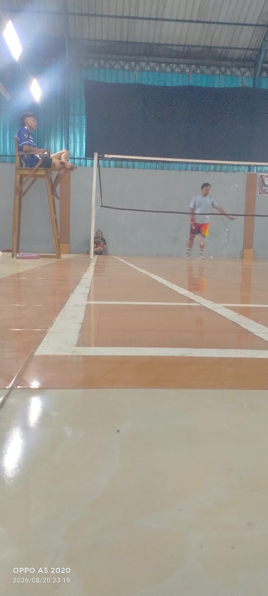 The image depicts an indoor badminton court in Dongos, Indonesia. In the foreground, the polished wooden floor of the court is visible, marked with white lines. In the background, a badminton net stretches across the court. To the left, a person sits on a raised wooden chair, appearing to be a referee or umpire. Further back, near the net, two individuals are present: one is seated on the floor, and another is standing on the court, holding a shuttlecock and appearing to be engaged in play or preparing to serve. The court is illuminated by overhead lights, and the walls are a neutral color, with a dark curtain or backdrop visible behind the net. The overall scene suggests a sporting event or practice session taking place within a covered facility.