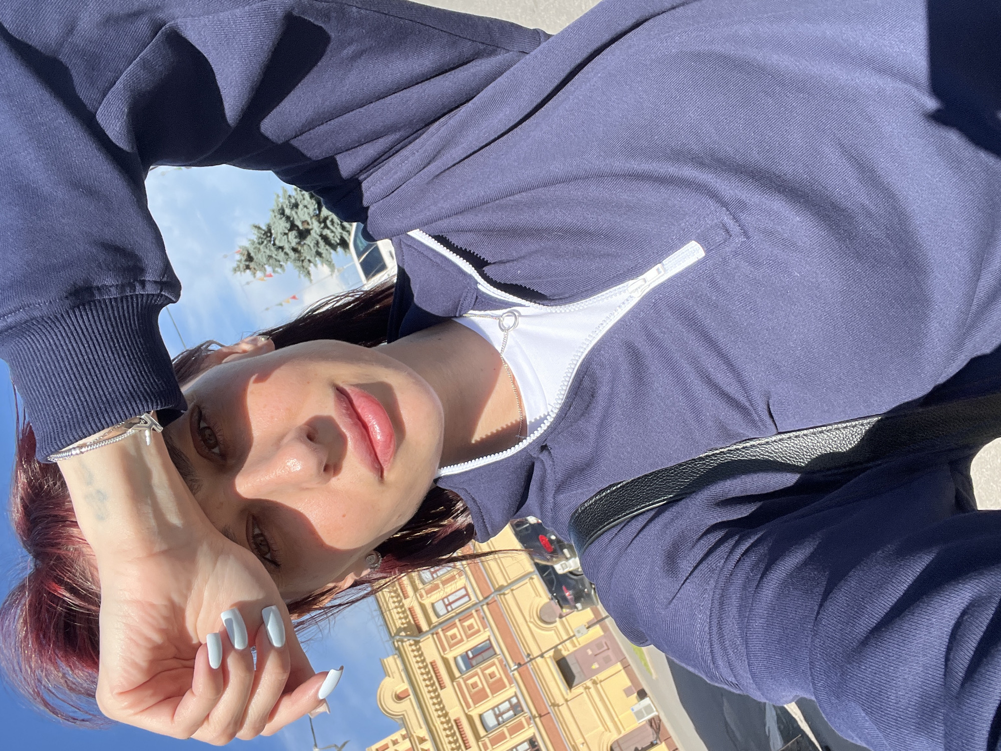 A young woman with dark red hair, light blue nail polish, and a bracelet on her wrist is taking a selfie outdoors. She is wearing a navy blue quarter-zip sweatshirt over a white t-shirt and a silver necklace. Her left hand is raised to shield her eyes from the sun. In the background, a yellow building with red accents and arched windows, a green coniferous tree, and a street with parked cars can be seen. The image appears to have been taken in Nizhny Novgorod, Russia, based on the architectural style of the building. The lighting suggests it is a sunny day.