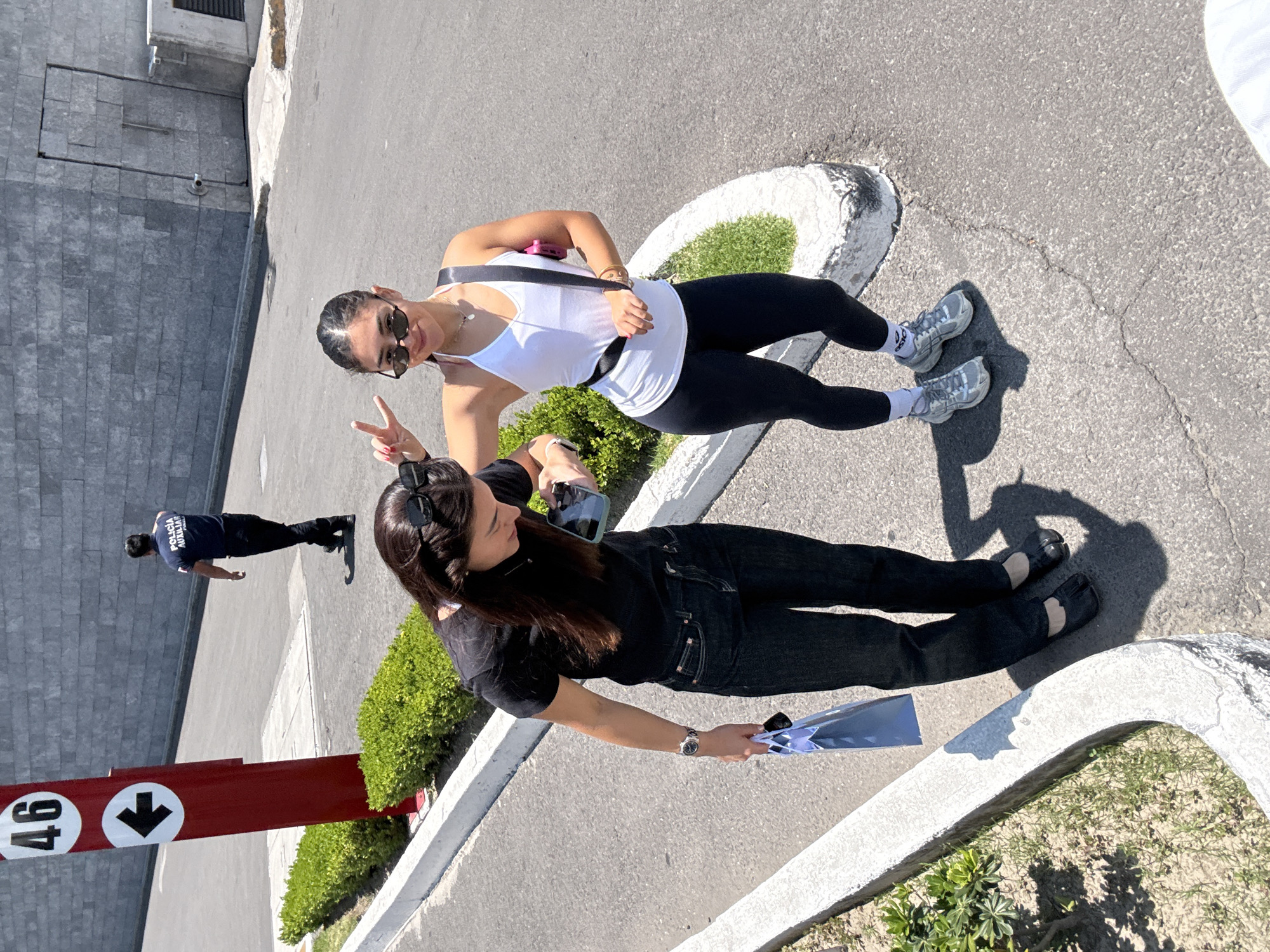 The image depicts an outdoor scene on a bright, sunny day, likely in Puebla, Mexico, given the provided context. Two women are posing for the camera on a paved surface, surrounded by strong shadows indicating clear skies and direct sunlight.
The woman closer to the viewer wears a black short-sleeved top, black pants, and distinctive black split-toe (tabi-style) shoes with light-colored footies. Her dark hair is pulled back, with sunglasses resting on her head. She holds a light blue or silver bag in her right hand and a smartphone in her left, looking towards the camera with a subtle smile. She wears a watch on her left wrist.
Behind her, the second woman is dressed in a white tank top, black leggings, and gray sneakers with white socks. Her dark hair is also pulled back, and she wears sunglasses, a necklace, and possibly bracelets. A black strap, likely from a crossbody bag or fanny pack, is visible with a small pink item attached. She is making a peace sign with her left hand and smiling cheerfully at the camera.
In the background, a man walks away from the camera. He wears a dark blue shirt with the white text "POLICÍA AUXILIAR" (Auxiliary Police) visible on his back. To the left, a prominent red post features two white circular signs: one with the number "46" and another with a downward-pointing arrow, possibly indicating a gate or specific location. The setting includes concrete dividers outlining patches of green shrubbery and a large wall constructed of gray, stacked blocks. A small security camera is visible on the wall. The overall atmosphere is light and cheerful.