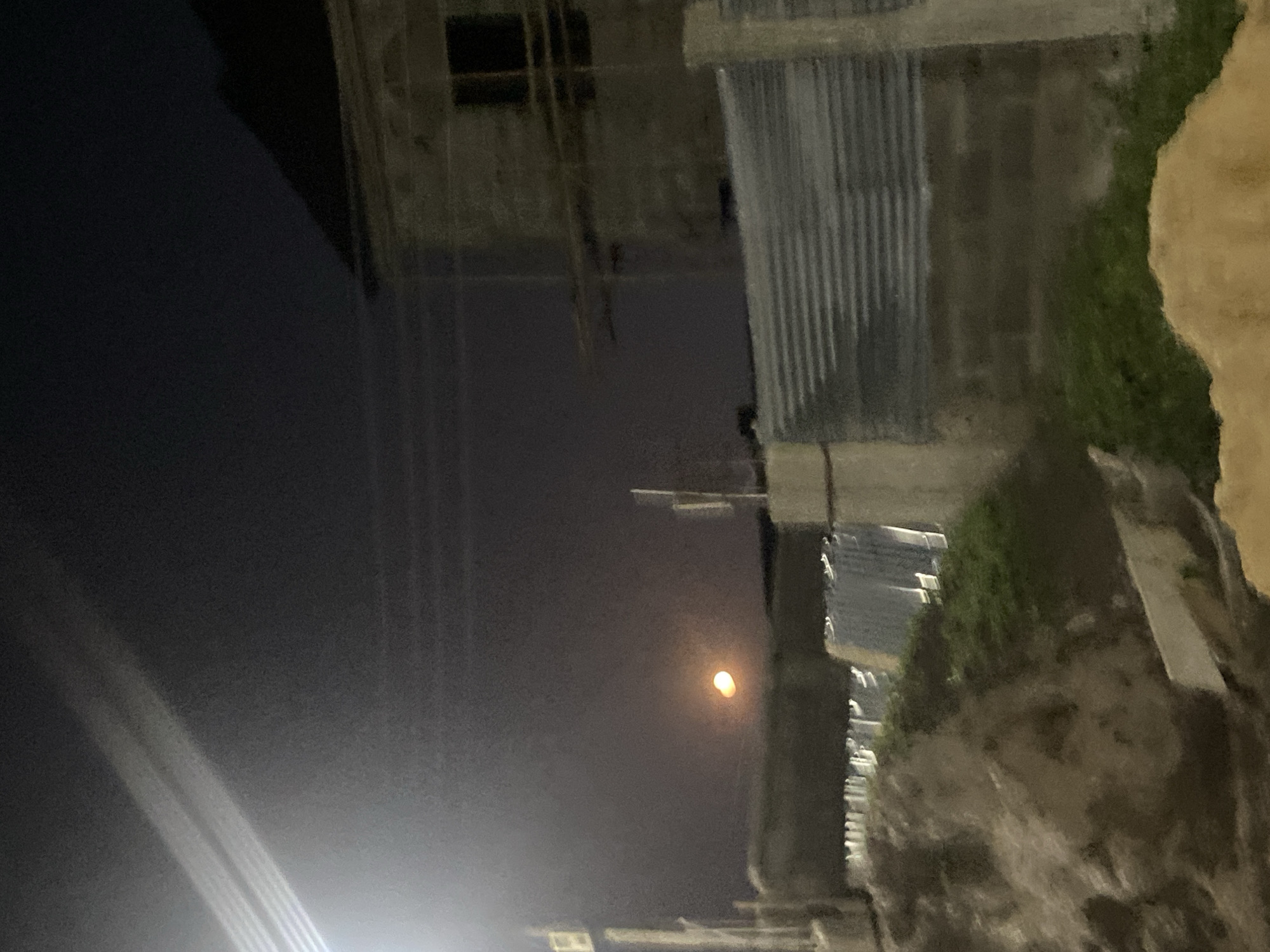 The image depicts a night scene in Lagos, Nigeria. A full moon illuminates the dark sky, visible between buildings. The buildings appear to be made of concrete blocks and corrugated metal sheets. There is some vegetation growing on the sides of some buildings. There are also electrical wires overhead.