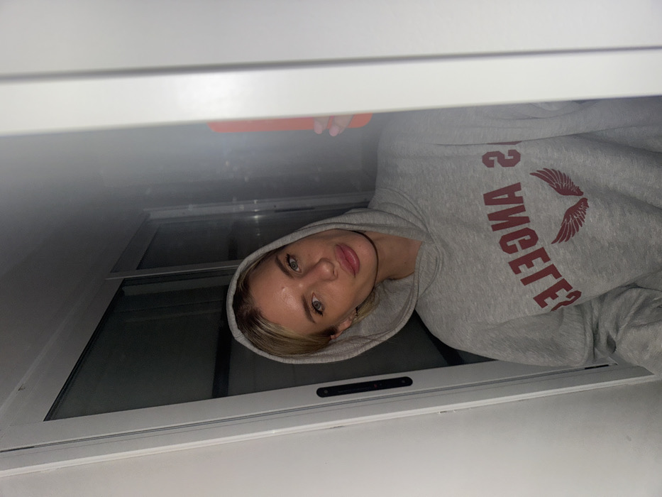 A young woman with light hair and fair skin wearing a gray hoodie with the words "LOS ANGELES" printed in red, along with a wing graphic, is peering down at the camera. She has green eyes and light pink lips. The shot is taken at an odd angle, framing her within the confines of a white window frame. A glimpse of an orange object is visible above her, possibly a phone or tablet. The scene is dimly lit, casting shadows that add depth to the composition.