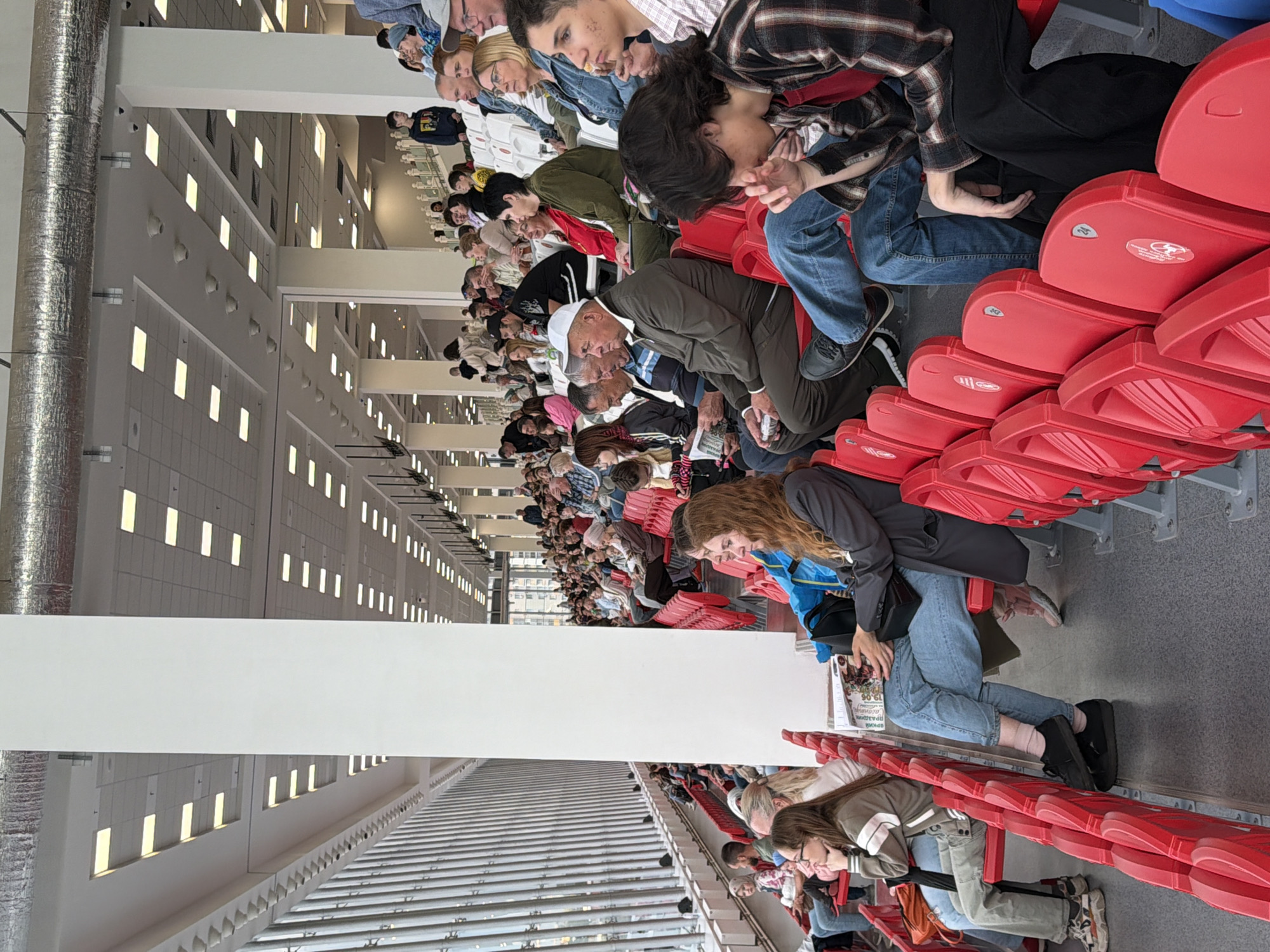 An indoor stadium with rows of red seats is filled with spectators. The ceiling is lined with fluorescent lights and large ventilation ducts. The architecture features tall white pillars and a long wall of vertical glass panels on the left side of the image. People of various ages are seated, some looking forward, others engaged in conversation, and some are partially obscured by the seats and pillars. The overall scene depicts a crowd attending an event in a large, modern indoor venue.