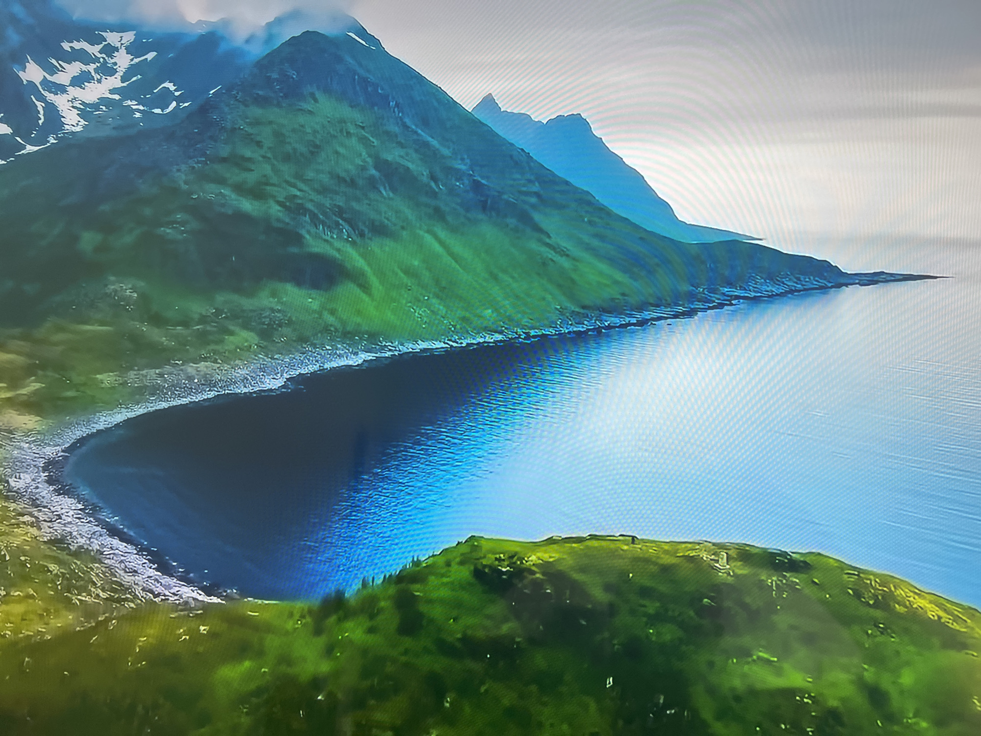 The image shows a coastal landscape with mountains. The mountains are green with some snow on the higher peaks. A body of water, likely the sea, extends into a bay. The water is blue with reflections. The coastline is rocky. There is green vegetation in the foreground. The sky is overcast.
The location in the city of Basingstoke and Deane, United Kingdom is not correct for this image. The terrain is mountainous. The city of Basingstoke and Deane has no coastline or mountainous regions.