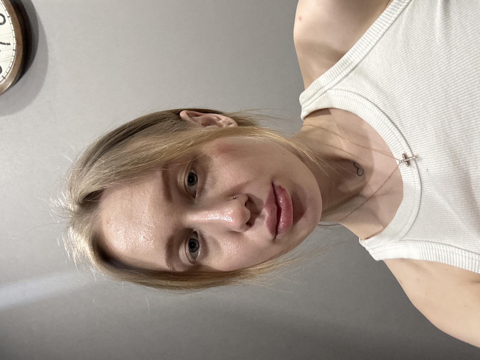 A single female subject is depicted in a close-up, upward-angled indoor portrait taken in Rostov-on-Don, Russia. The subject has light-colored hair, styled loosely around her face, and light-colored eyes. Her complexion is fair, with a subtle sheen visible on her forehead, nose, and chin, and a slight pinkish flush on her right cheekbone. A small metallic stud piercing is present in her left nostril. She wears a white ribbed tank top. A rose gold-colored chain necklace with a small, jewel-encrusted cross pendant rests on her sternum. A small black outline tattoo, depicting a heart, is visible on the left side of her neck/upper chest area. A small red mark is present on her upper right shoulder. The background consists of a plain, light grey wall. A portion of a round, wooden-framed wall clock with black numerals is partially visible in the upper right corner of the image. The subject is looking directly into the camera with a neutral expression.