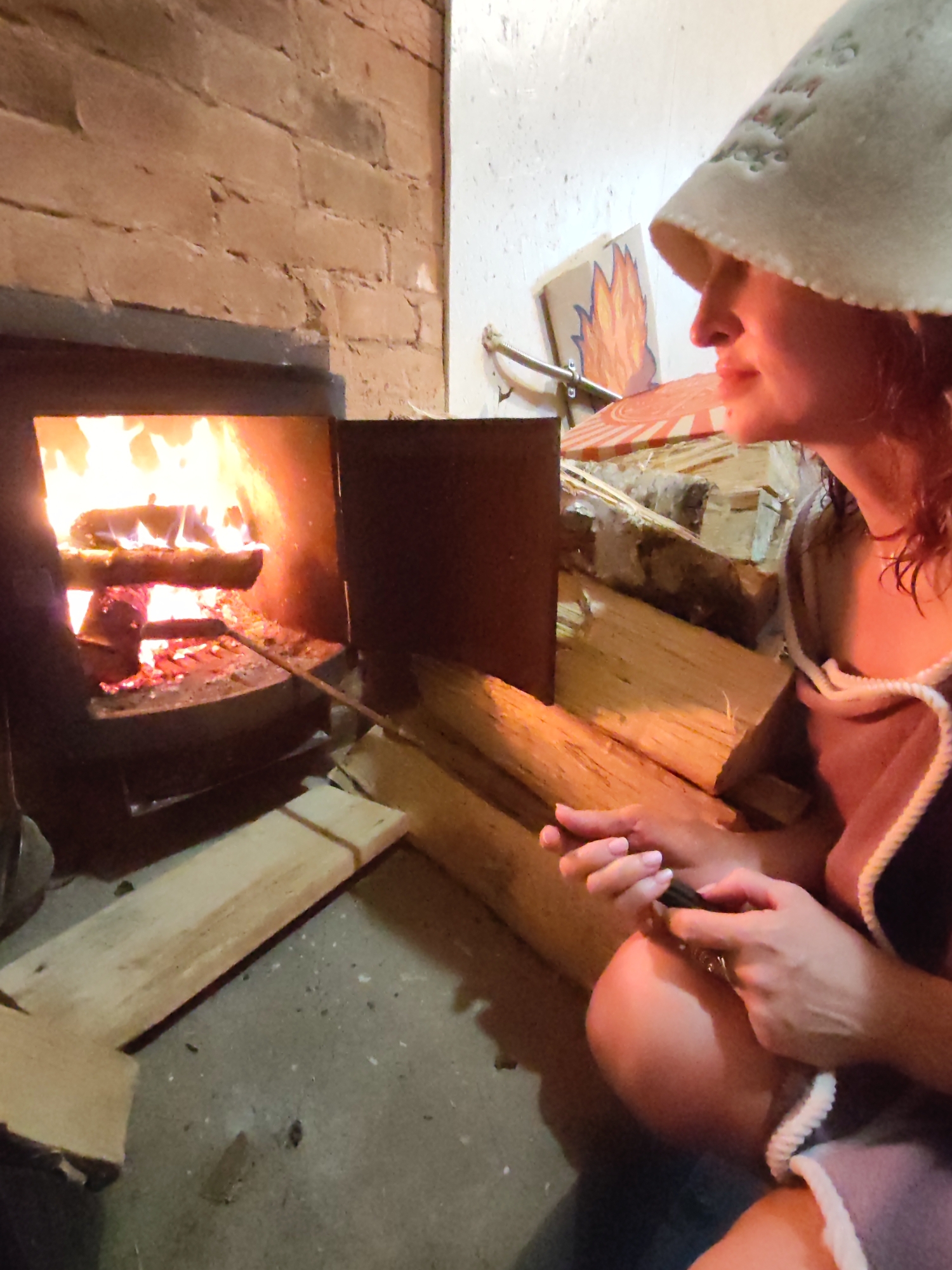 A woman wearing a felt sauna hat and a robe is sitting near a brick fireplace. She is holding a metal tool in her hands. A fire is burning inside the fireplace, and there is wood stacked next to it. The location is Unknown, Unknown.