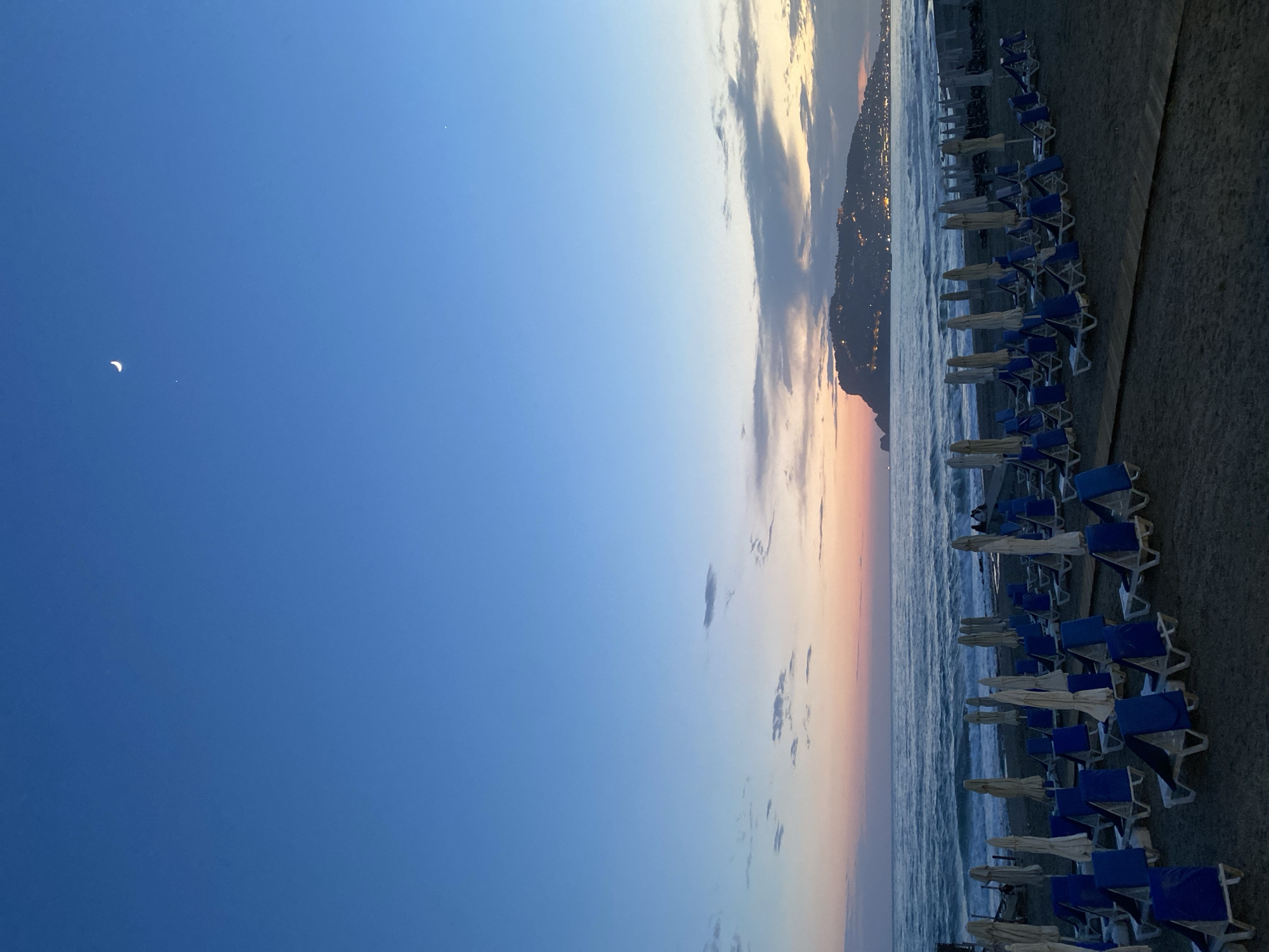 The image depicts a beach scene in Alanya, Turkey, at dusk. A crescent moon is visible in the upper left corner of the dark blue sky. The sky transitions to lighter shades of blue and orange near the horizon, where clouds are present. The Mediterranean Sea meets the beach, with gentle waves visible. A row of beach chairs and closed umbrellas line the shoreline. In the background, a silhouette of a mountain is present, with city lights visible along the coast, indicating the location as Alanya. There are no people visible in the scene.