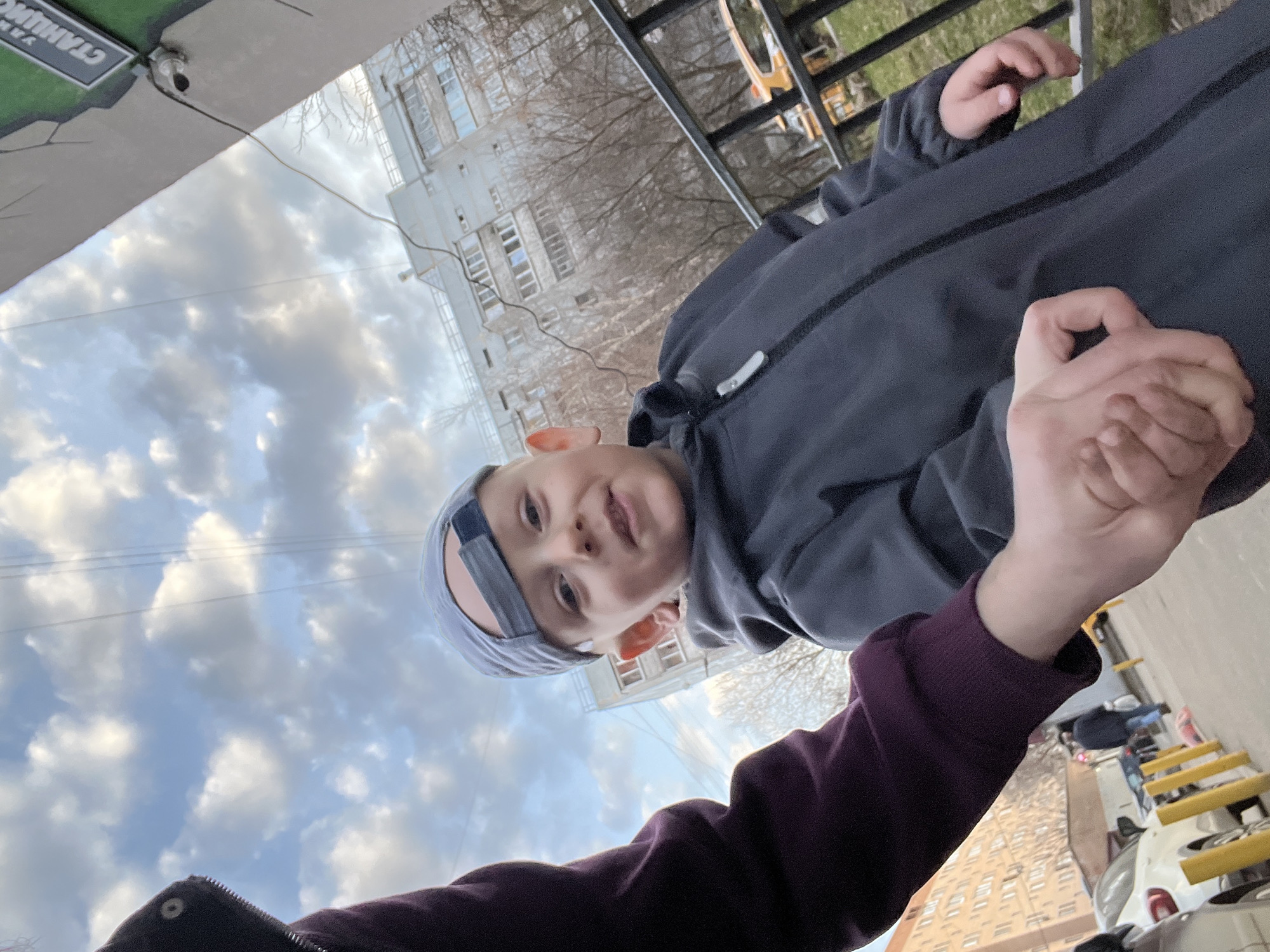 A low-angle shot shows a child in a grey tracksuit and blue bandana looking at the camera. Their hand is clasped by an adult, whose purple-sleeved arm is also visible. The background shows a residential building, cloudy sky, a balcony with railings, and street parking in Mytishchi, Russia.