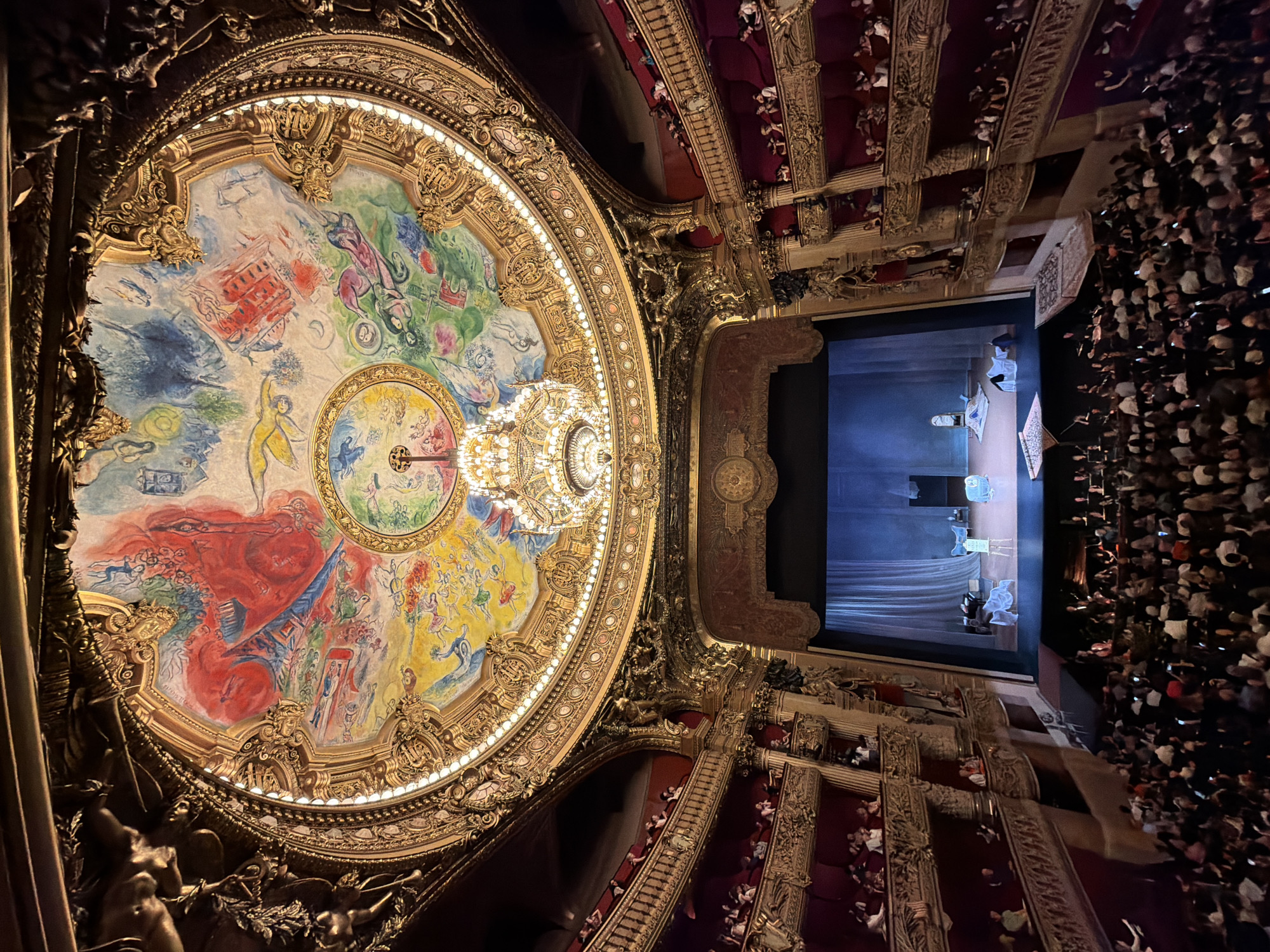 The image captures the ornate interior of the Palais Garnier opera house in Paris, France, from an elevated perspective. The focal point is the Chagall ceiling, a large circular painting adorned with vivid colors and whimsical figures. A grand chandelier hangs from the center of the ceiling, adding to the opulence of the space. Below, the theater's stage is visible, with actors performing under a blue light. Rows of seats are filled with spectators, extending upwards and towards the edges of the frame. The overall impression is one of grandeur and artistic splendor, reflecting the historical and cultural significance of the venue.