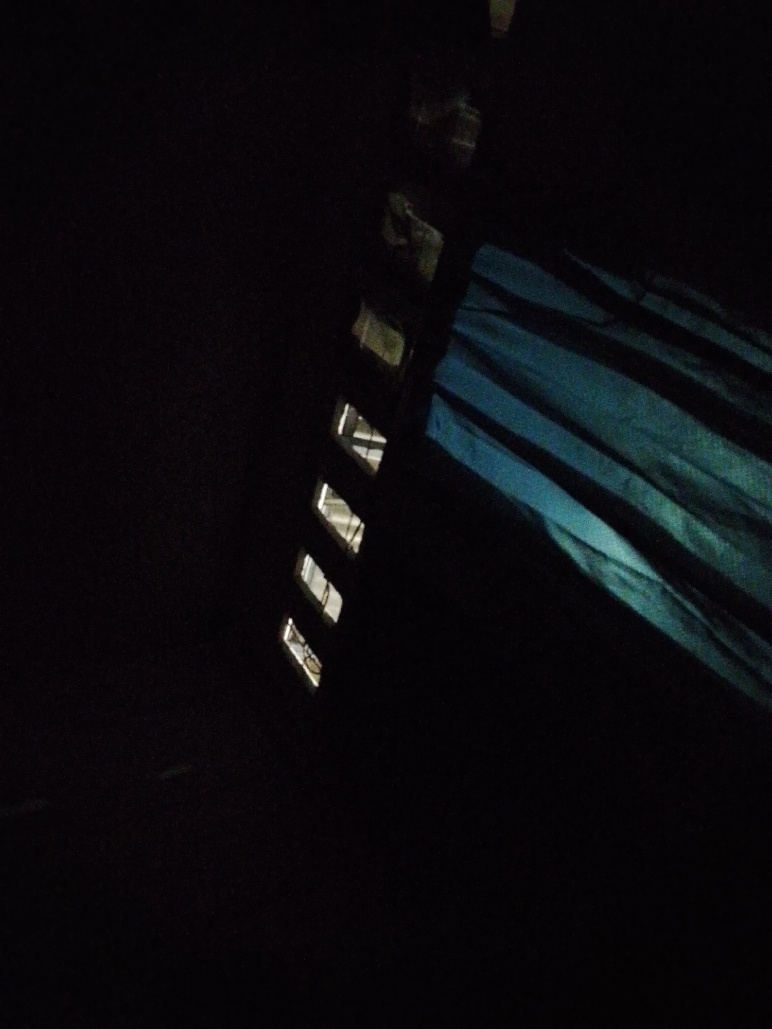 The image depicts an exterior view of a building in Bukit Ketok, Indonesia, at night. Light emanates from rectangular openings, possibly windows, along a vertical facade on the right side of the frame. The remainder of the image is predominantly dark, obscuring detail. A blue drape or curtain is visible on the right side.