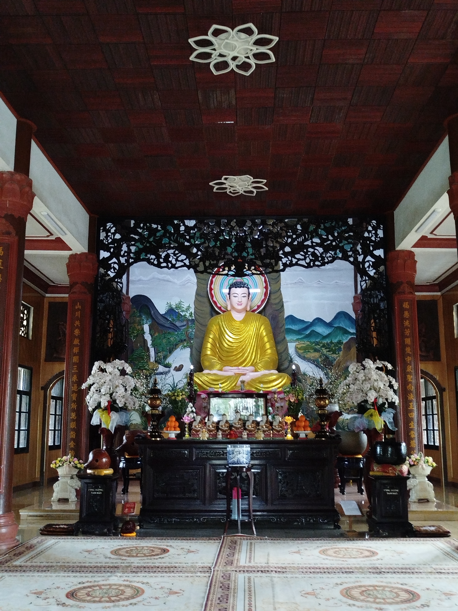 The image shows the interior of a temple in Xuân Trường - Đà Lạt, Vietnam. A golden Buddha statue sits in a meditative pose behind a dark wooden table that is decorated with candles, incense, and food offerings. The statue is set against a painted backdrop of mountains and trees. Flanking the table and Buddha statue are vases of white flowers. Tall, dark wooden columns, some of which are inscribed with golden text, support a dark wooden ceiling with geometric patterns and decorative floral lighting fixtures. The floor is covered with a patterned carpet. Windows are visible on the left and right, allowing natural light to enter the space.