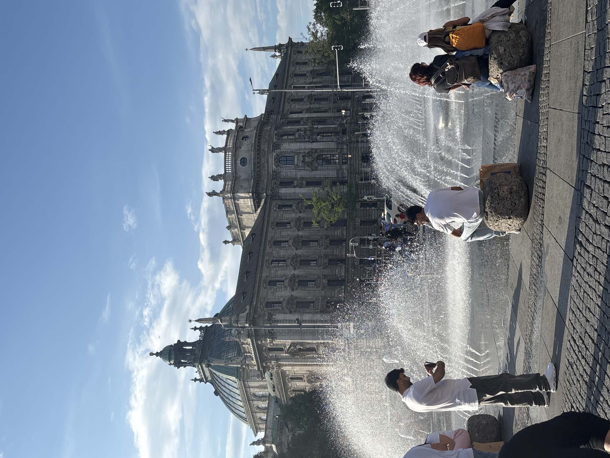 The image shows a view of the Fountain of the Four Rivers (Vierbrunnnenbau) in front of the Palace of Justice (Justizpalast) in Munich, Germany. The Palace of Justice is a grand, ornate building with a large dome and classical architecture. In the foreground, a large fountain sprays water, creating a shimmering effect in the sunlight. Several people are seated on large, rough-hewn stones near the fountain, some looking at their phones, others simply observing the surroundings. A man in a white shirt and grey pants stands and looks at his phone, while a few people sit on stones, facing away from the camera, with backpacks. The sky is blue with scattered white clouds. The scene depicts a public space in a city, likely a tourist attraction, with people enjoying a sunny day by the fountain and the impressive architecture.