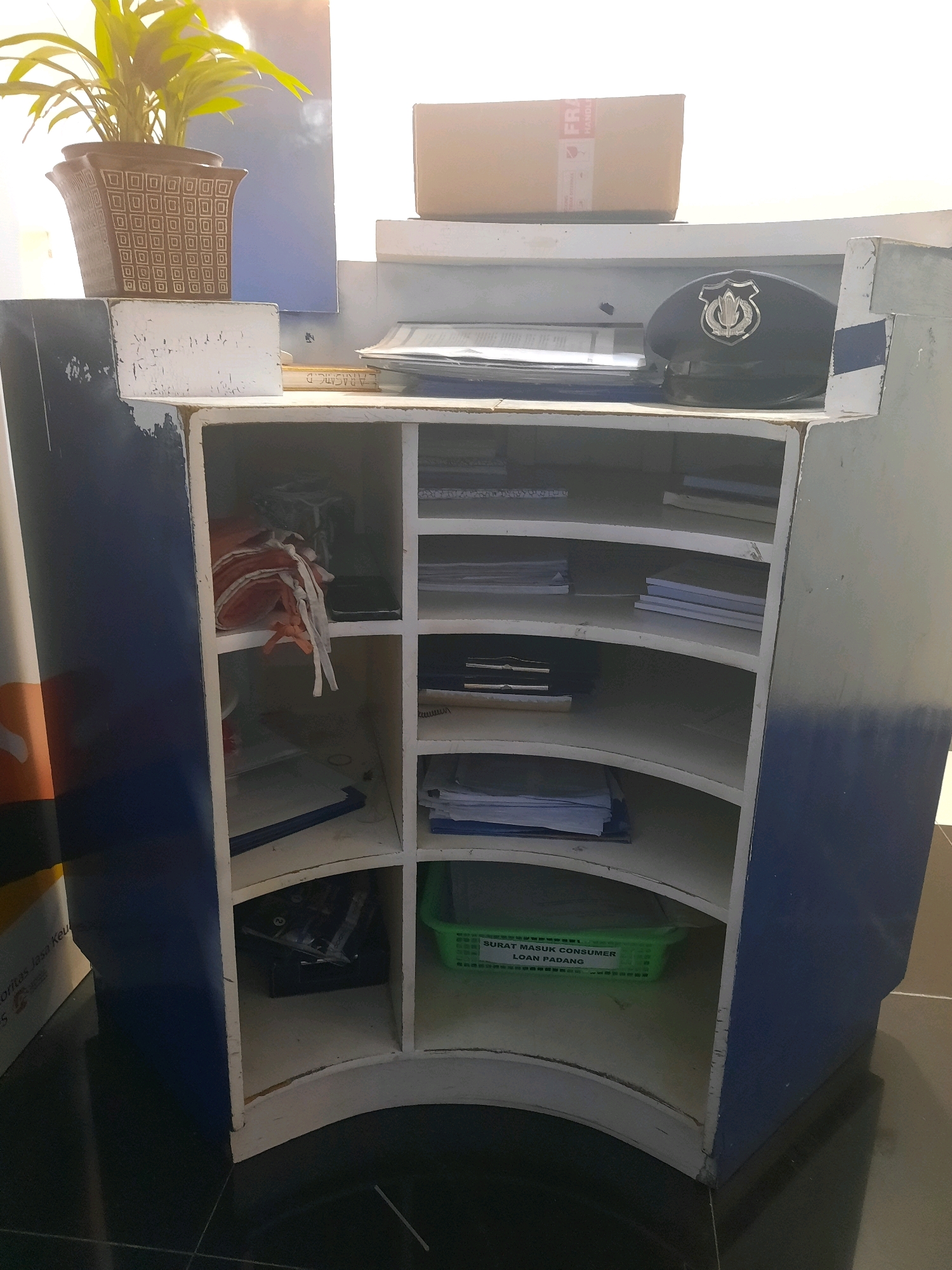 The image shows a cluttered reception desk at what appears to be a loan office in Padang, Indonesia. A plant in a decorative pot sits on top of the desk, next to a stack of papers and a dark blue cap with a silver emblem. The desk itself is divided into several compartments, some of which are filled with papers, folders, and other office supplies. One compartment contains a green plastic basket labeled "SURAT MASUK CONSUMER LOAN PADANG," indicating the contents are incoming mail for consumer loans in Padang. The desk is made of wood and is painted white and blue, with visible signs of wear and tear. The floor is dark and tiled. The overall impression is one of a functional but somewhat disorganized office space.