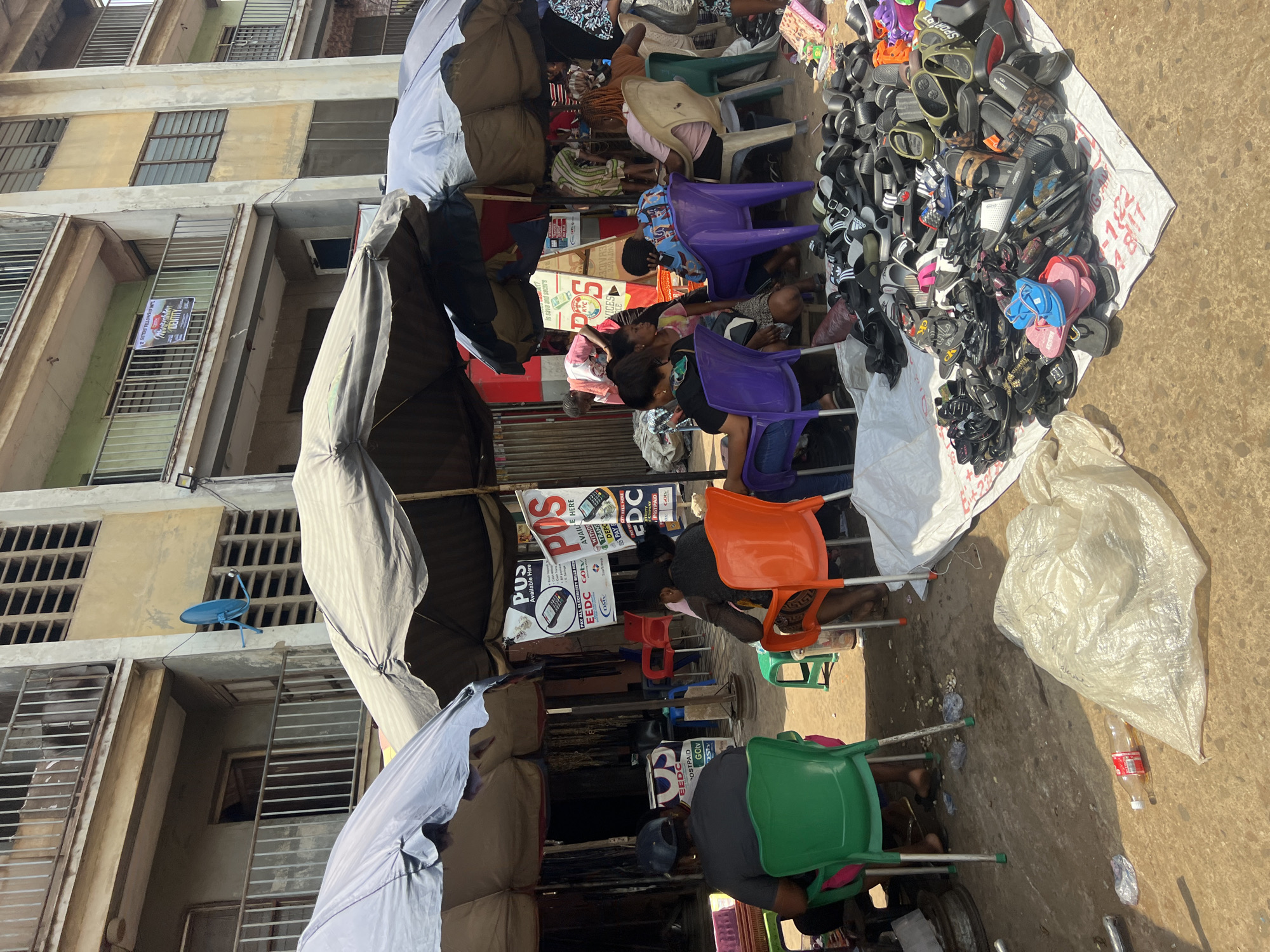 The image shows a bustling street market scene in Onitsha, Nigeria, characterized by its vibrant commercial activity. The foreground is dominated by a large assortment of footwear displayed on a white sheet. Customers are seated on an assortment of plastic chairs of varying colors (purple, orange, green) while interacting with vendors. There are signs advertising services such as POS (Point of Sale) availability and payment of electricity bills. These signs include EEDC and Gotv logos. An open umbrella provides shade for the vendors and their wares. The background features a multi-story building with barred windows and fading paint, common architecture in urban commercial areas of Nigeria.