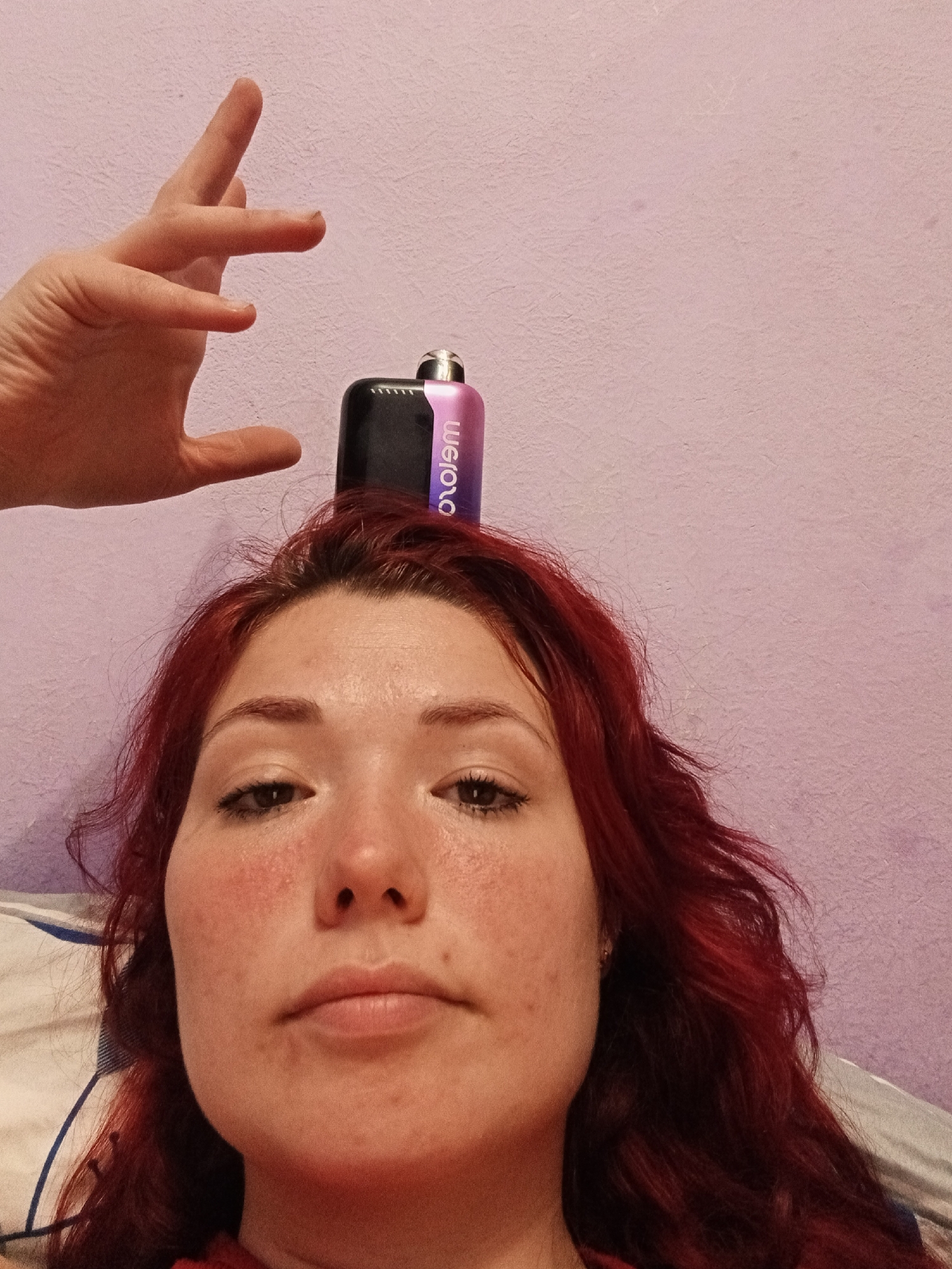 An overhead shot captures a woman with vibrant red hair resting on a pillow, her face prominently featured in the foreground. A black and purple vape device is balanced on her head, with a hand extended from the top left of the frame, fingers poised just above the device, as if about to adjust it. The background is a textured, pale pink wall. The woman's complexion is fair with visible blush and freckles. She is looking directly at the camera. This image was taken in Chaikovsky, Russia.