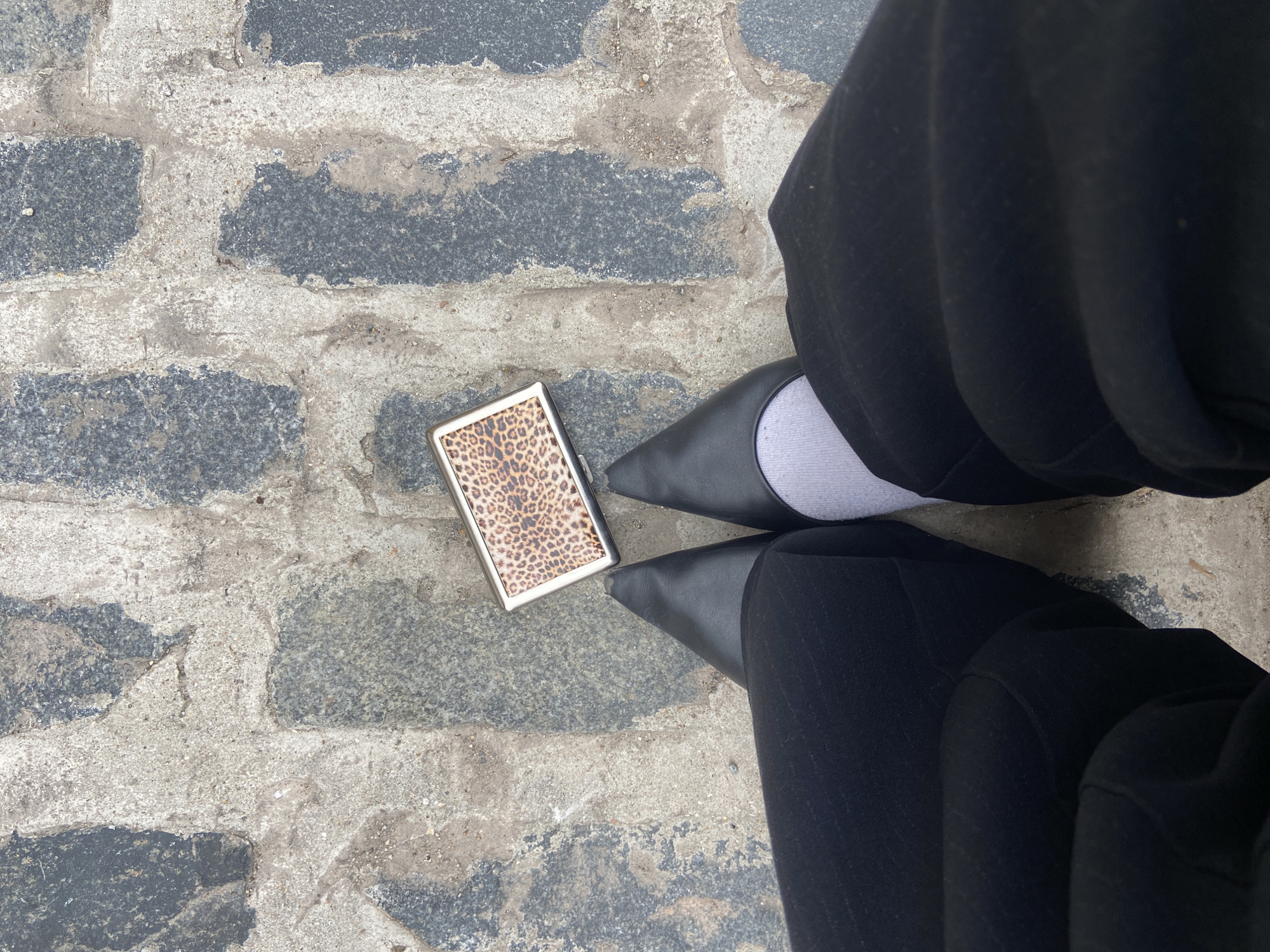 The image displays an overhead view of an individual's lower legs and feet positioned on a paved surface in London, United Kingdom. The ground is composed of irregular dark grey stone blocks set within light grey, worn mortar, with some minor debris visible. The individual wears dark-colored trousers, black pointed-toe flat shoes, and a light grey sock visible above the right shoe. Centered directly in front of the feet, a small, rectangular object with a metallic silver-colored frame and a prominent leopard print pattern on its upper surface rests flat on the pavement.