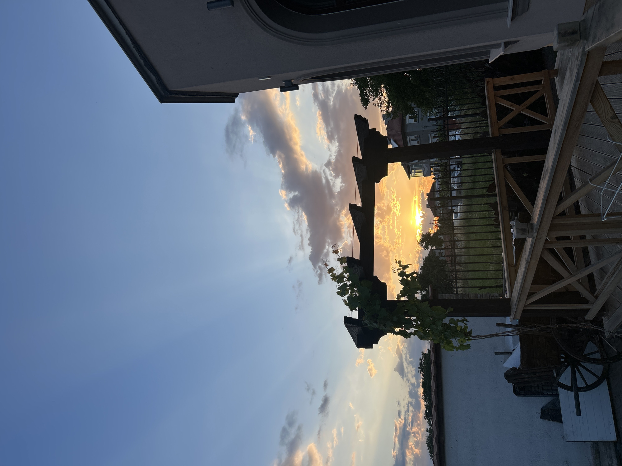 The image captures a sunset scene in Voyvodino, Bulgaria. The sky is filled with dramatic clouds illuminated by the setting sun, casting a warm golden and orange glow. Rays of light pierce through the clouds, creating a beautiful atmospheric effect.
In the foreground, a wooden pergola with dark wooden beams and a vine growing on it stands. To the right, a wooden deck and railing are visible, along with part of a building's white wall and some outdoor furniture, including an antique wagon wheel. Behind the pergola, a metal fence encloses a grassy area. In the distance, several buildings are visible, suggesting a residential area.
The time of day is clearly sunset, indicated by the low sun and the colors in the sky. The weather appears clear with scattered clouds. The scene evokes a sense of calm and natural beauty. There is no visible text in the image.