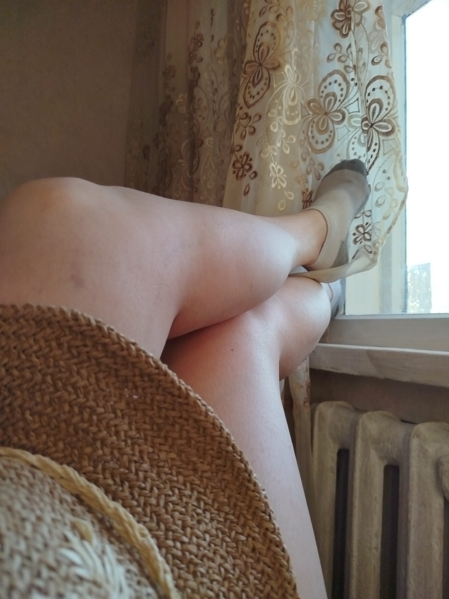 A person's legs are visible, with one leg crossed over the other. The person is wearing light-colored socks and is partially covered by a woven straw hat. The scene appears to be indoors, with a curtain patterned with flowers and a radiator visible in the background. The lighting suggests it is daytime. There is no indication of the location within the city of Qarağandı city administration, Kazakhstan.