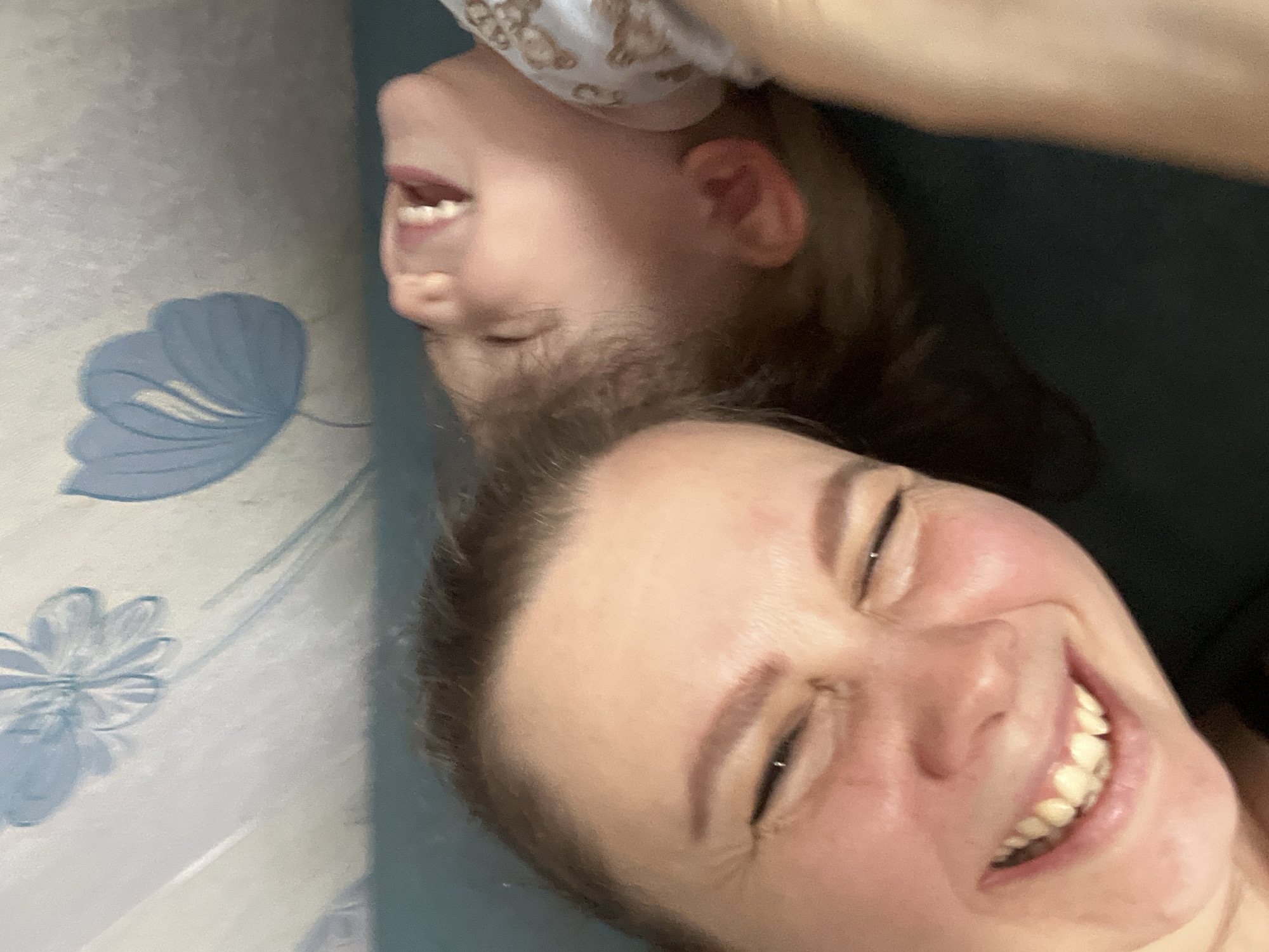 A selfie taken indoors shows a woman and a child. The woman is in the foreground with a broad smile, revealing her teeth. Above her, oriented upside down, is a child with an open mouth, also possibly smiling. The background is a mix of light blue wall with a floral pattern and a dark blue surface. The location is unknown.