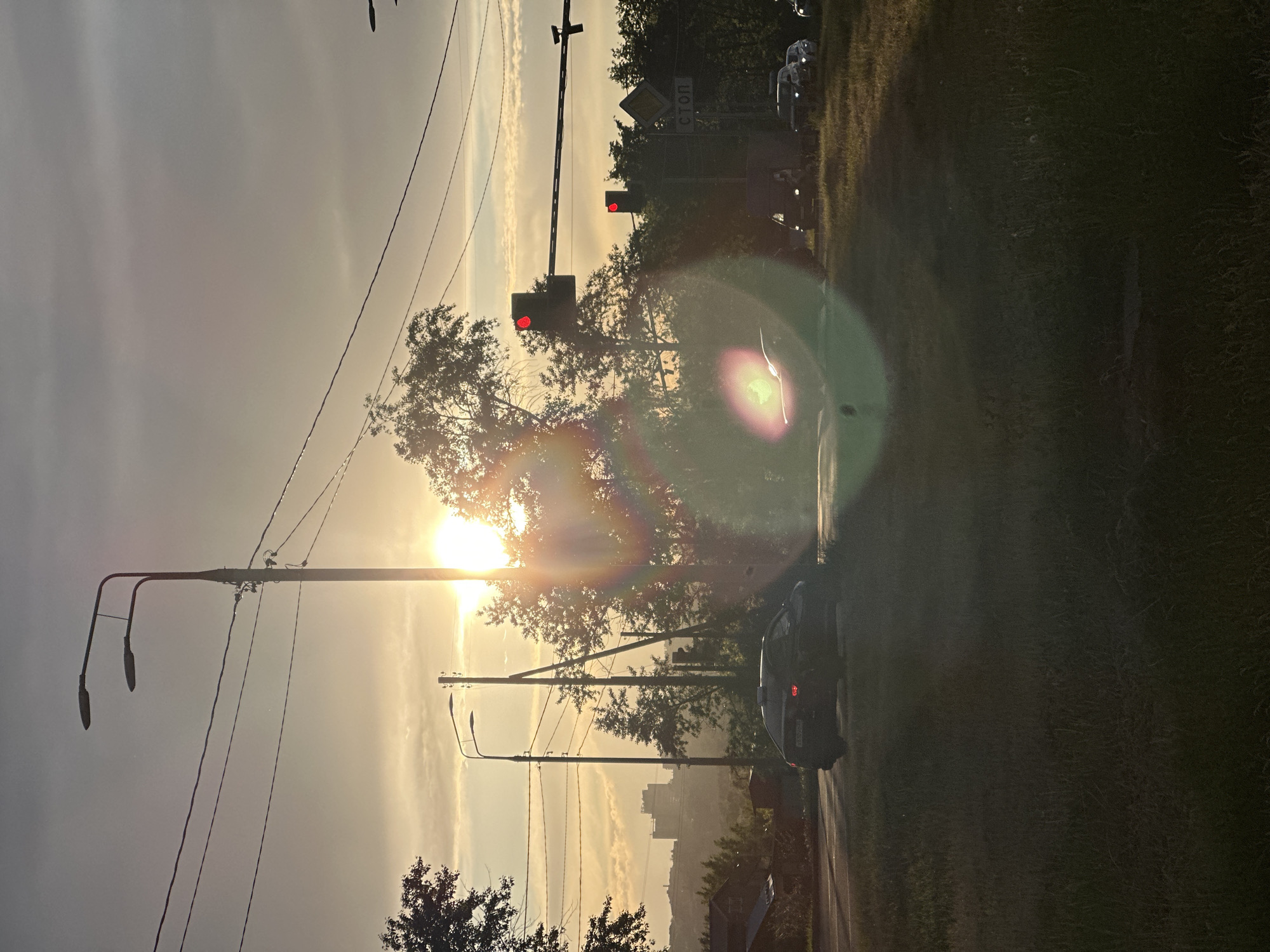 The image shows a street scene in what appears to be Vladimir, Russia, taken during a golden hour. The sun, low in the sky, is causing a lens flare. A car with its taillights illuminated is driving on the right side of the road. Traffic lights are visible, with both sets displaying red lights. Power lines and streetlights are present. There's vegetation alongside the road. In the distance, the silhouette of buildings hints at the city's urban layout. A "СТОП" sign is visible near the traffic lights.