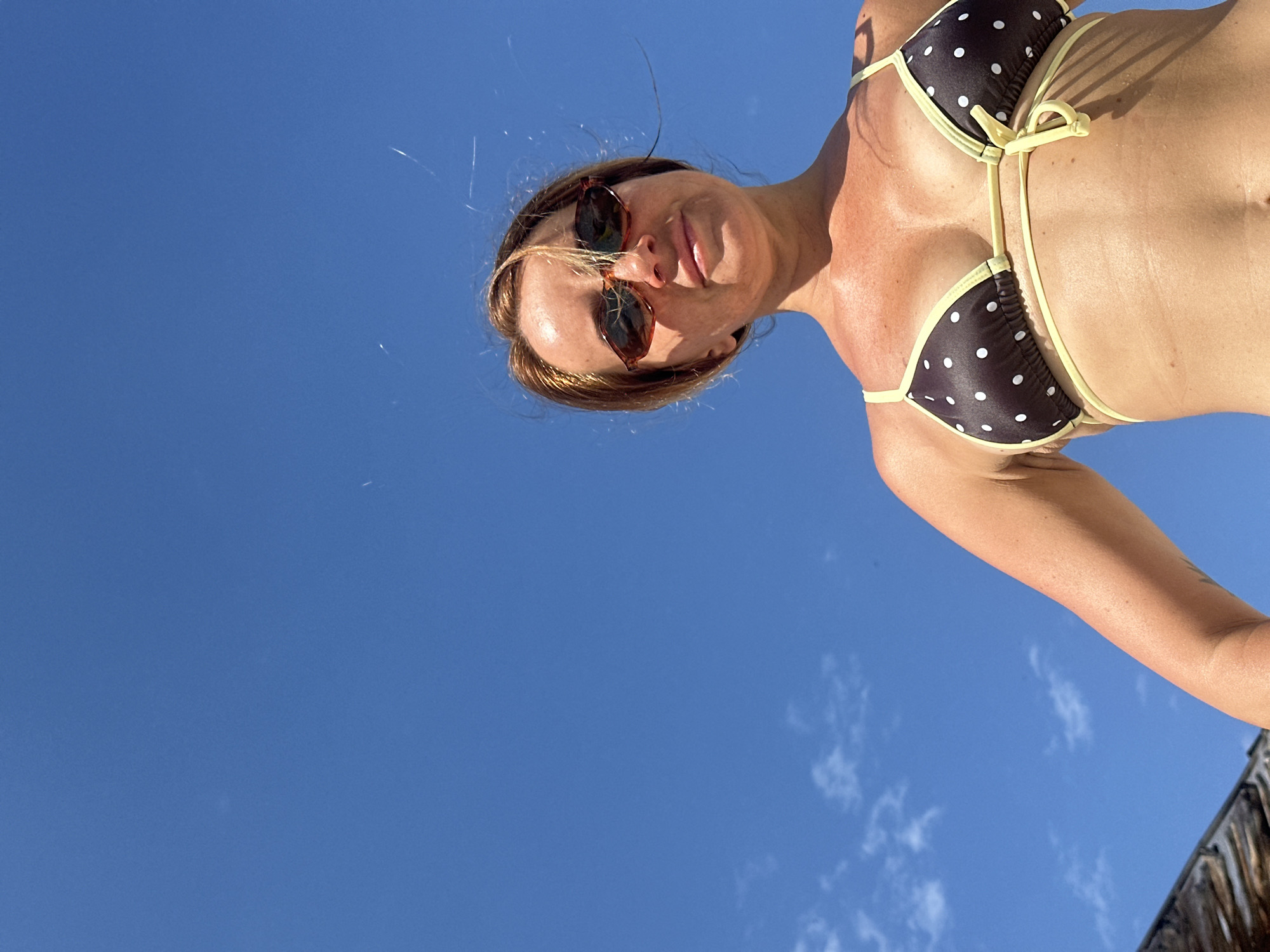 An outdoor, low-angle, waist-up shot features a woman in a brown and white polka dot bikini against a bright blue sky with a few wisps of white clouds. She is wearing brown sunglasses and her hair is a light brown. Her arms are raised slightly, and she is looking at the camera. The bottom left of the frame shows a sliver of a straw-thatched structure. The scene appears to be a beach in North Kuta, Indonesia.