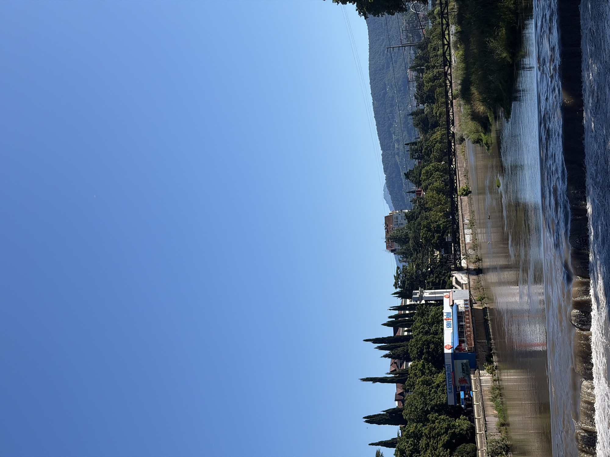 The image depicts a wide, calm river flowing through a city, likely Sochi, Russia, under a clear blue sky. In the foreground, a low waterfall or weir on the river creates whitewater. Along the riverbank, there is lush green vegetation and several buildings, including a two-story establishment with "Santorini" and "Start Up" signage. Tall, slender trees, possibly cypress, line the bank in front of residential buildings with red-tiled roofs. A pedestrian bridge with a metal railing crosses the river in the middle ground. Beyond the city buildings and trees, a heavily wooded mountain range rises in the background, with some power lines visible. The overall scene suggests a tranquil, sunny day in a city situated near mountains and a river.