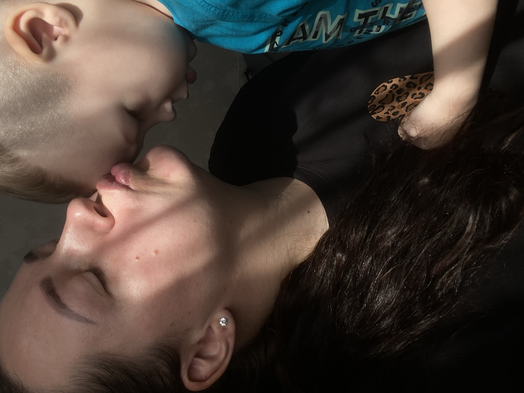 A close-up shot depicts a mother and child in intimate contact. The mother lies on her back with her eyes closed. She wears a black shirt with a leopard print patch and a diamond stud earring in her left ear. Dark hair spreads around her head. The child, wearing a blue shirt, appears to be kissing the mother's lips. The setting is not discernible, and there is no evidence to suggest that the image was taken in Sochi, Russia.