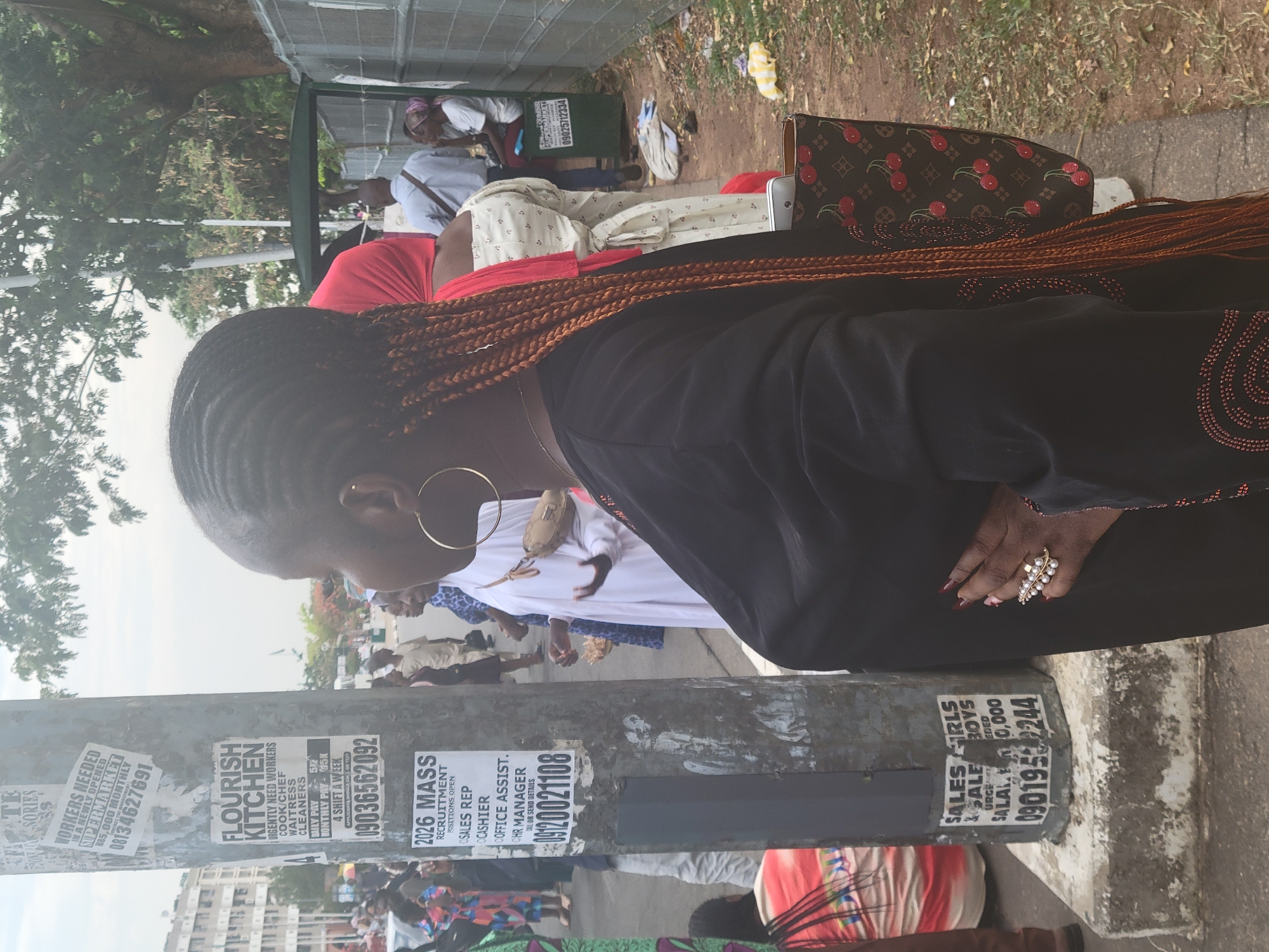 The image depicts a street scene in Abuja, Nigeria. A woman with braided hair stands near a utility pole covered in various job advertisement posters. The posters advertise positions at businesses like Flourish Kitchen, 2026 Mass Recruitment, and a supermarket.
In the background, other people are visible, including individuals seated under a tent-like structure and others standing and walking. The woman is wearing a dark-colored dress with beaded embellishments. She has a gold hoop earring and a ring with pearls.
