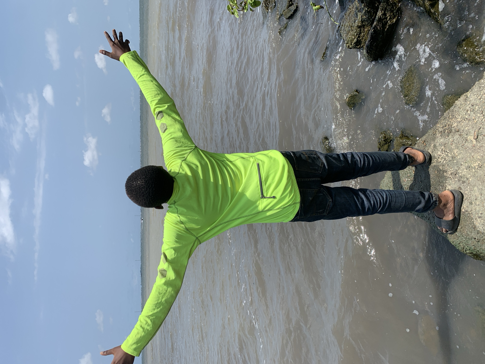 The image depicts a person standing by the edge of a body of water, potentially a river or lagoon, in what appears to be Ikorodu, Nigeria. The individual, seen from the back, wears a neon green jacket and dark jeans. They are standing on a concrete bank with one foot on a grassy area near the water's edge, and their arms are outstretched. The water is murky, and there are rocks and vegetation along the shoreline. The sky is partly cloudy.