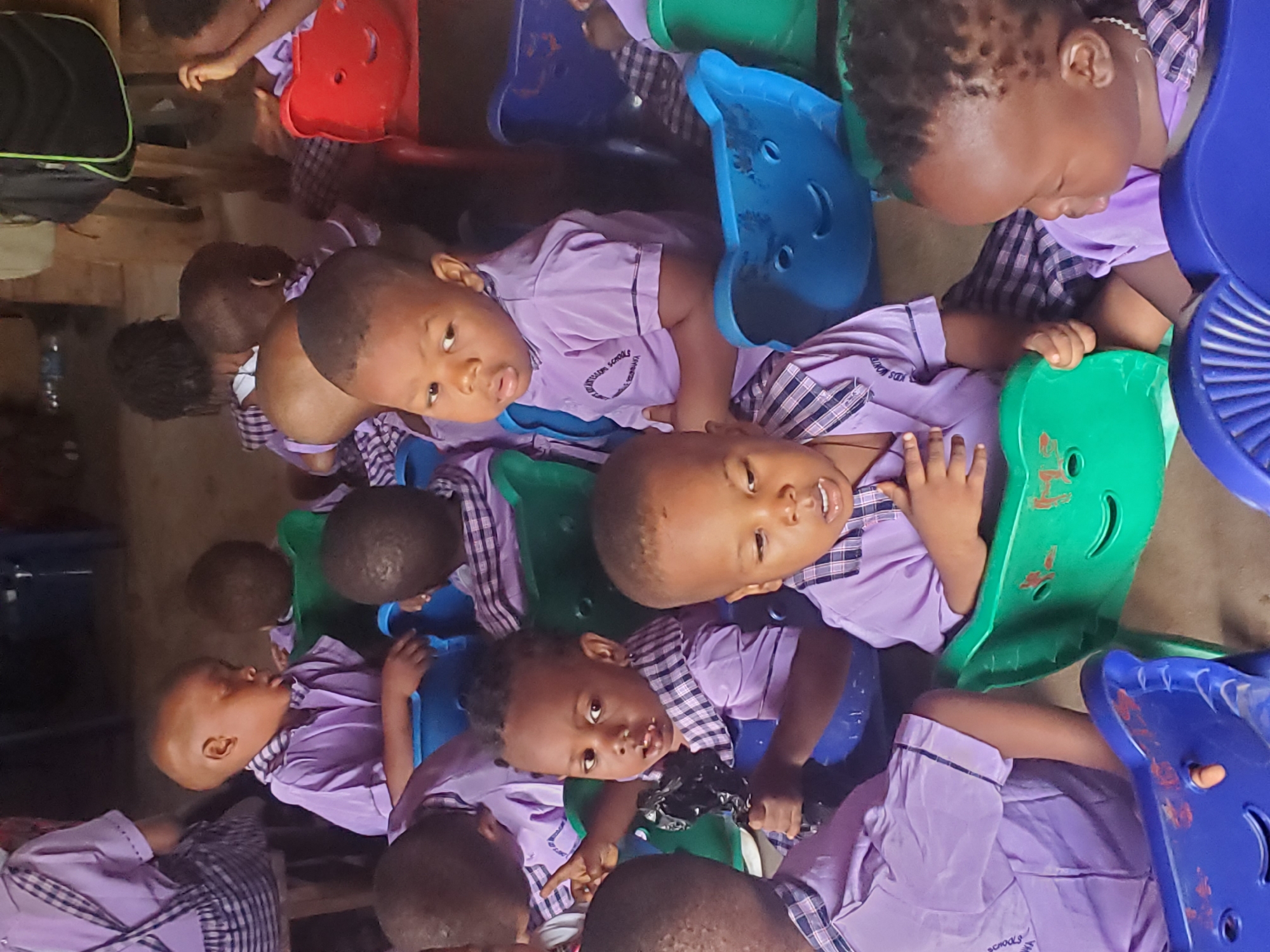 The image shows a group of young children, presumably in a classroom setting in Ukwu-Abwa, Nigeria. They are seated in colorful plastic chairs (blue, green, red), each wearing a light purple short-sleeved uniform shirt with a checkered collar. The shirts have the name of the school on them, Kid's Mountain School. The children are of various skin tones. Some are looking towards the camera, while others are looking down or away. The room has a simple, unadorned appearance. A black backpack is visible in the upper left corner of the image.