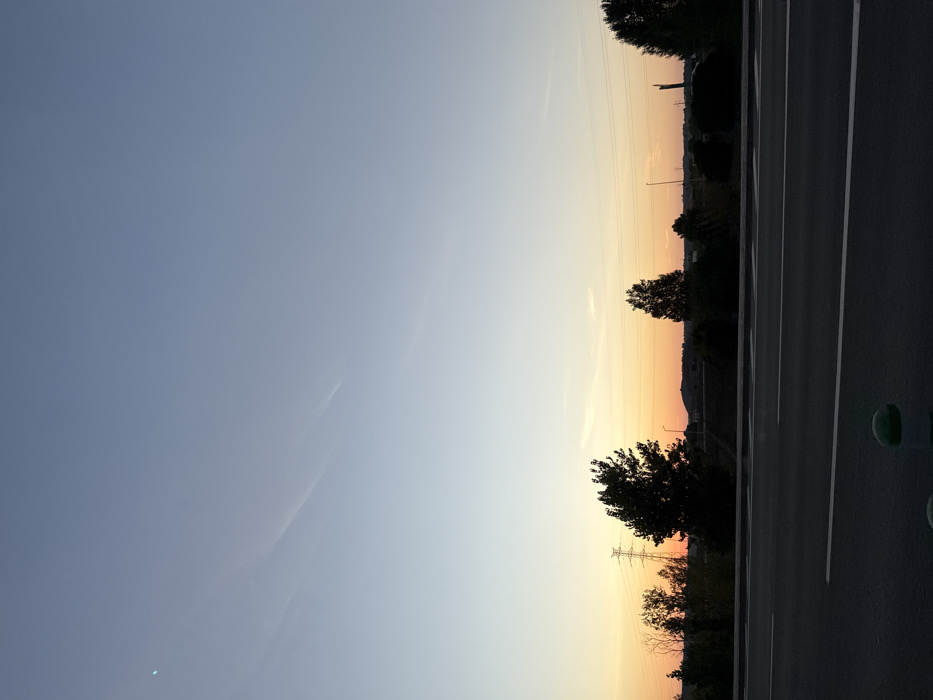 This media file captures a tranquil landscape scene, most likely during either sunrise or sunset, with the location context being Santarém, Portugal. The image is dominated by a vast, clear sky that presents a beautiful color gradient. It transitions from a soft blue-grey at the top to warm shades of orange and yellow closer to the horizon, indicative of dawn or dusk. Faint, wispy clouds or contrails are subtly visible in the upper portions of the sky, and a very small, crescent-like white speck, possibly the moon, can be seen in the upper left corner.
Along the right side of the frame, a dark asphalt road stretches into the distance, marked with clear white lines that delineate lanes or shoulders. Several green, circular objects, possibly bollards or road markers, are spaced along the immediate edge of the road.
The horizon is defined by the dark silhouettes of numerous trees and a distant, undulating landscape, which could be hills or the outline of a distant town. Overlaid against the vibrant sky, slender power lines and utility poles are visible, hinting at human infrastructure blending into the natural environment. No people or visible text are present in the image. The overall atmosphere conveyed is one of peace and natural beauty under a transitioning sky.