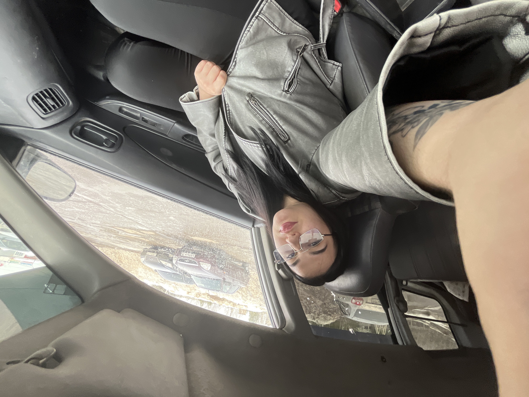 A female, approximately 20-35 years old, with dark hair and rectangular metal-framed glasses, is seated in the front passenger seat of a vehicle, capturing a selfie. She wears a grey zippered faux-leather jacket and dark clothing; a tattoo depicting foliage or feathers is visible on her left forearm. Her right hand is positioned on the zipper of her jacket.
The vehicle's interior features dark upholstery, with the front passenger door panel displaying window controls, a door handle, and an air vent.
Outside the vehicle, visible through the windows, is a wet, unpaved, or partially snow-covered ground surface, indicating melting conditions or recent precipitation. Several vehicles are parked, including a dark red sedan with Russian license plate E694EP 18 (Perm Krai region 18), a grey hatchback with Russian license plate ending in 159 (Perm Krai region), and another dark sedan with Russian license plate 265YY 159 (Perm Krai region). In the background, a snow-covered embankment with sparse, bare trees is visible, along with a distant building.
This scene is located in Chaikovsky, Perm Krai, Russia, as evidenced by the region codes on the visible Russian license plates. The overall environment suggests late winter or early spring conditions.