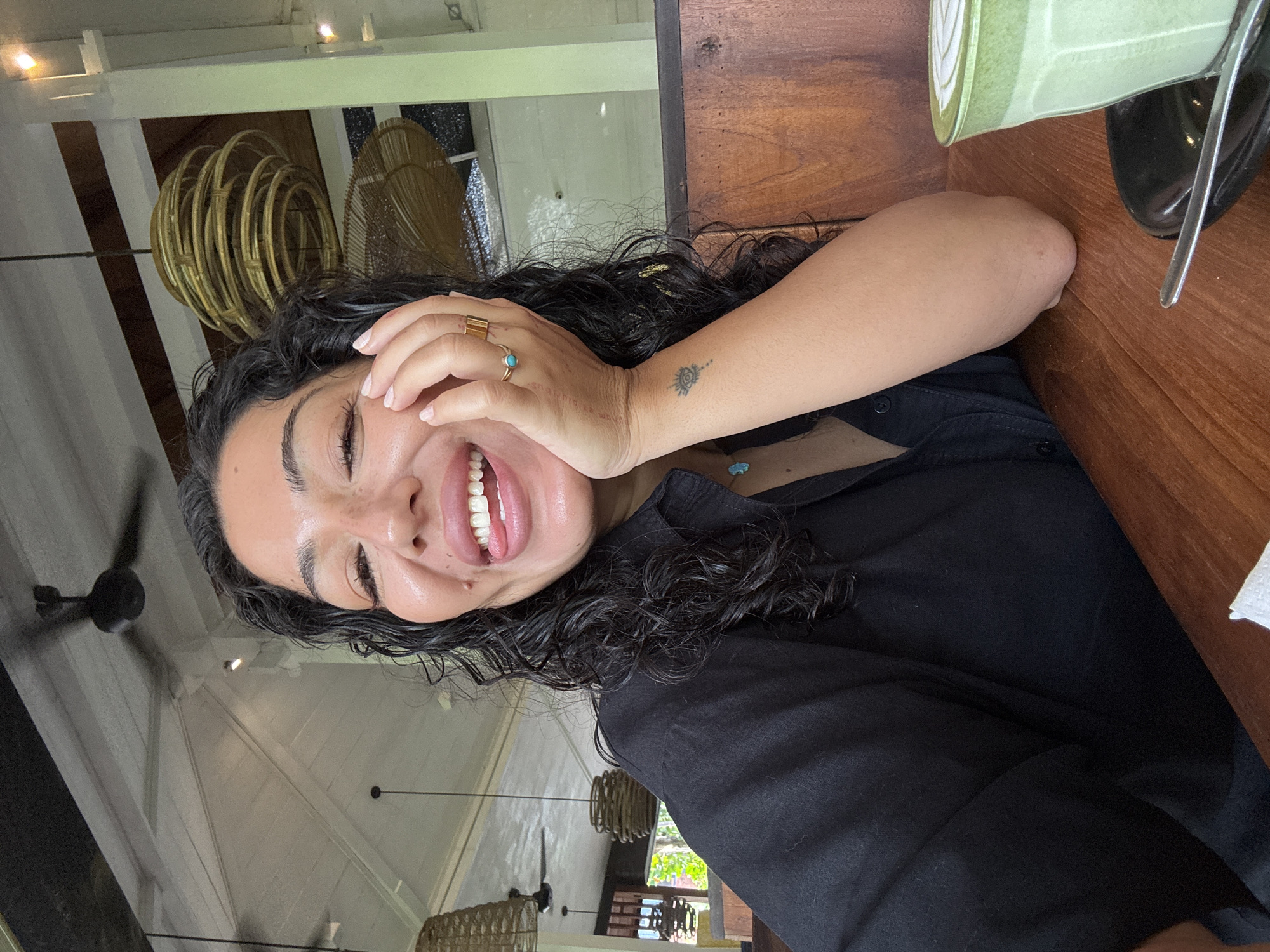 A close-up, eye-level photograph captures a person with dark, curly hair and fair skin, smiling broadly with closed eyes, conveying strong emotion like laughter or joy. The subject's right hand rests on her head, adorned with a simple gold band ring and another gold ring featuring a small, light blue stone. A small, stylized tattoo, possibly an abstract sun or eye, is visible on her left forearm. She is wearing a black collared shirt or top, with a hint of a light blue necklace or charm peeking out near her collarbone.
The image is taken from a slightly tilted perspective, placing the subject in the foreground on the left side. To her right, a warm-toned wooden table holds a light green cup, possibly containing a beverage like matcha, with a spoon or straw inside, and a dark bowl or dish.
The background reveals an indoor setting with elements suggesting an open-air or tropical environment, consistent with the North Kuta, Indonesia context. White-painted wooden ceiling panels are visible, along with multiple decorative light fixtures made from natural materials like wicker or rattan. One prominent spherical fixture hangs in the mid-ground, and a dark ceiling fan is also noticeable. The bright lighting suggests it is daytime. The overall mood of the image is joyful and relaxed. There is no visible text.