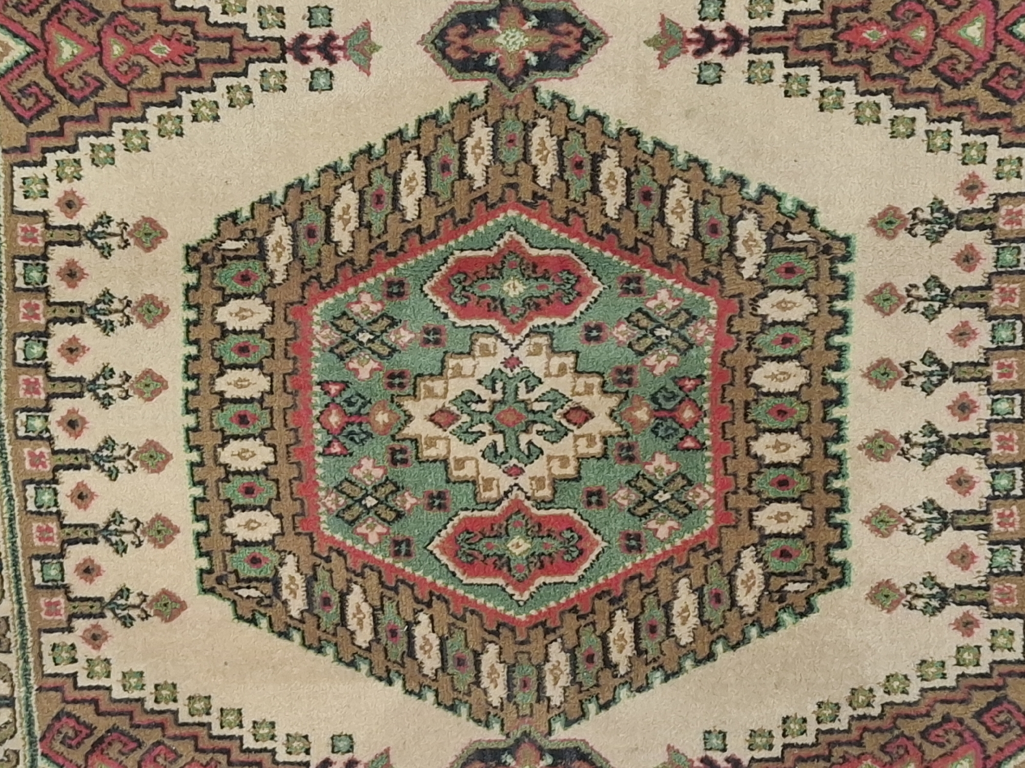 The image is a close-up, overhead shot of a section of a rug. The rug features intricate geometric patterns in shades of cream, brown, green, and red. A prominent hexagonal motif with floral and diamond-like elements is at the center. The surrounding areas have repeating patterns of smaller geometric shapes and what appear to be stylized floral or tree motifs. The texture of the rug is visible, suggesting it is made of woven material. There are no people, actions, or interactions depicted in the image. The environment is confined to the surface of the rug itself. There is no information in the image that can be used to determine its location in Almaty, Kazakhstan.