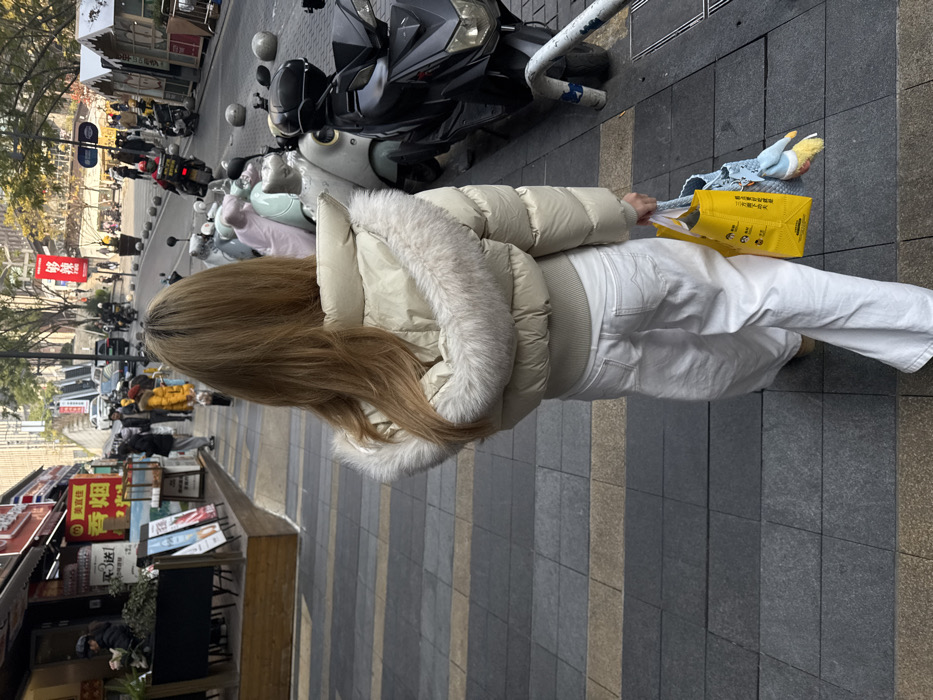 A woman with long, blonde hair is walking on a sidewalk beside a street with parked motorcycles. She wears a white puffer jacket with fur trim and white pants. She carries a yellow shopping bag in her right hand. Shops and street signs are visible in the background, along with pedestrians. The street appears to be in an urban setting during daylight.