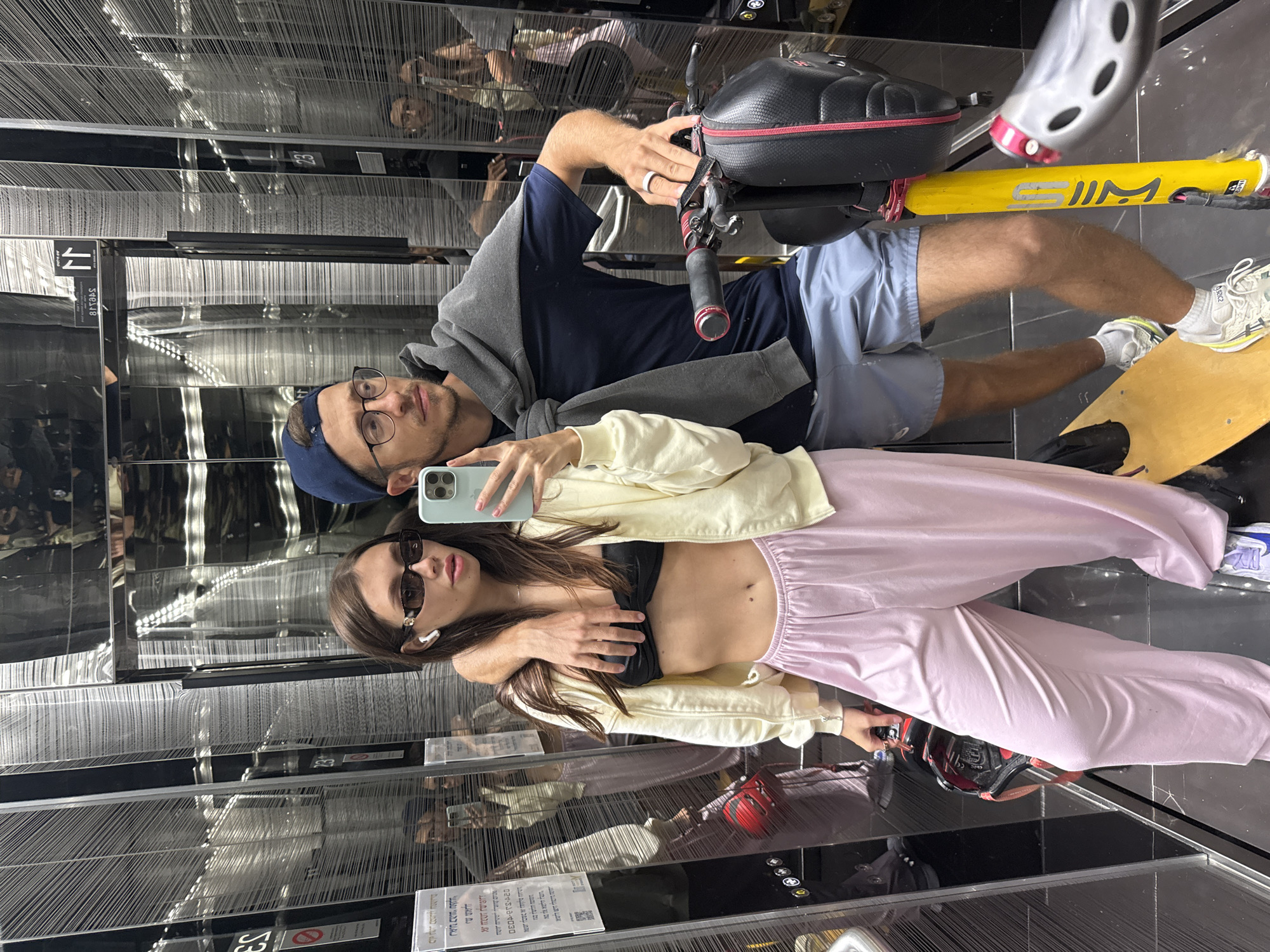 Two people are taking a selfie in an elevator. The elevator appears to be located in Tel-Aviv, Israel, based on the elevator signage in Hebrew and English. The male is wearing glasses, a blue cap, and a navy blue shirt with a grey sweater draped over his shoulders, and blue shorts. The female is wearing sunglasses, a black sports bra, a cream jacket, and pink sweatpants. A bicycle is positioned behind the couple. The elevator interior is primarily mirrored with linear patterned walls. The floor is black.