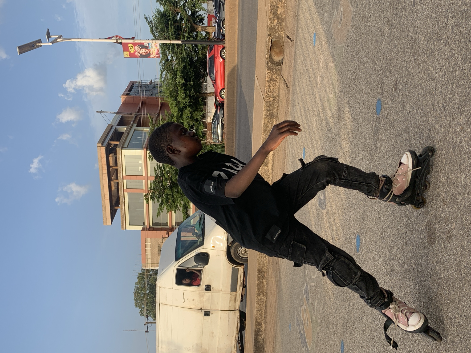 A youth is roller skating on a street in Jos, Nigeria. He is wearing black jeans, a black t-shirt with the word "VANROUGE" visible, and roller skates that appear to have pink tennis shoes attached. The location appears to be near a curb or roadside. A white van is parked nearby with a person visible in the driver's seat. In the background, there are buildings, trees, and a lamppost with a banner. Several blue markings are visible on the road.
