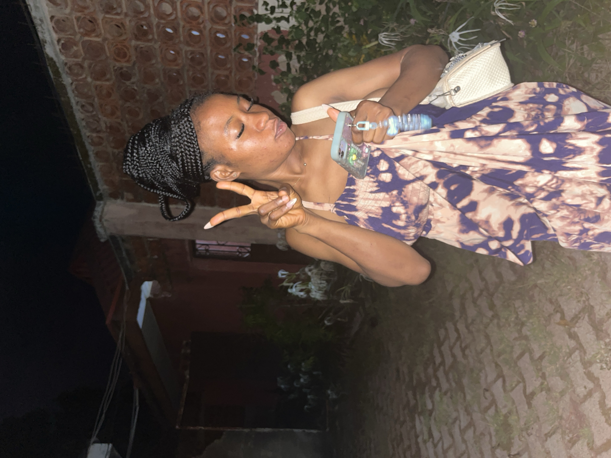 A young Black woman with her hair in braided extensions styled in a high bun is posing for a photo at night in a tiled outdoor area. She is wearing a sleeveless dress with a tie-dye pattern of purple and white. In her right hand, she holds a light blue smartphone, and in her left, she holds up two fingers in a peace sign. A white crossbody bag hangs from her right shoulder. The background features a brick wall and overgrown vegetation with white flowers. The scene appears to be a residential area, possibly in Jos, Nigeria, with a dark, unlit environment suggesting nighttime.
