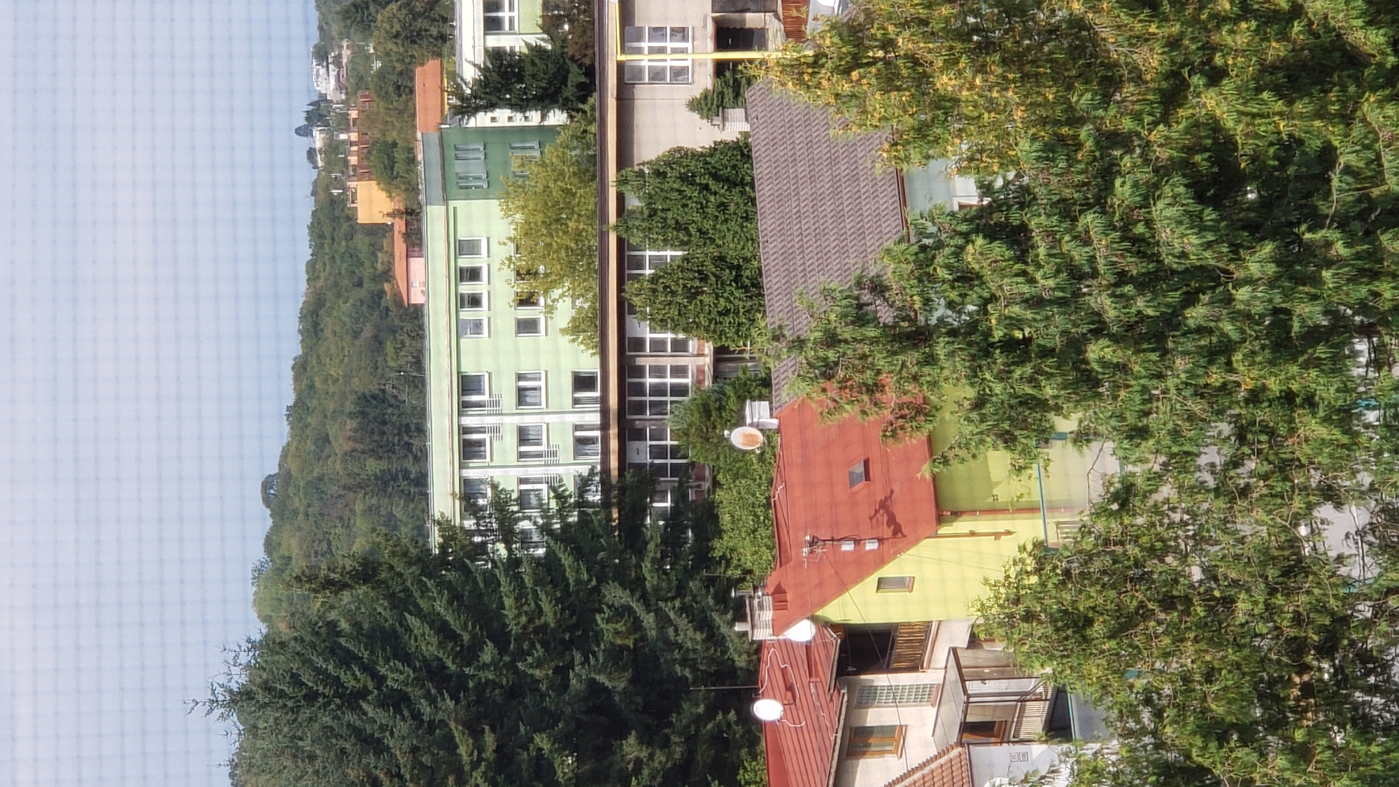 The image depicts a view of buildings and trees in the city of Prešov, Slovakia. In the foreground, there are several residential buildings with red and gray tiled roofs, interspersed with dense green trees. One building has a bright yellow facade and a red roof, while another adjacent building has a lighter colored facade and a gray tiled roof. Further back, a large, light green apartment building with multiple floors and many windows dominates the mid-ground. Behind this building, a grey, lower building with a long row of windows is visible. The background shows a hilly landscape covered with lush green trees, and in the far distance, several structures and buildings are scattered across the horizon, suggesting a suburban or city outskirts setting. The sky above is overcast. There are no people visible in the image.